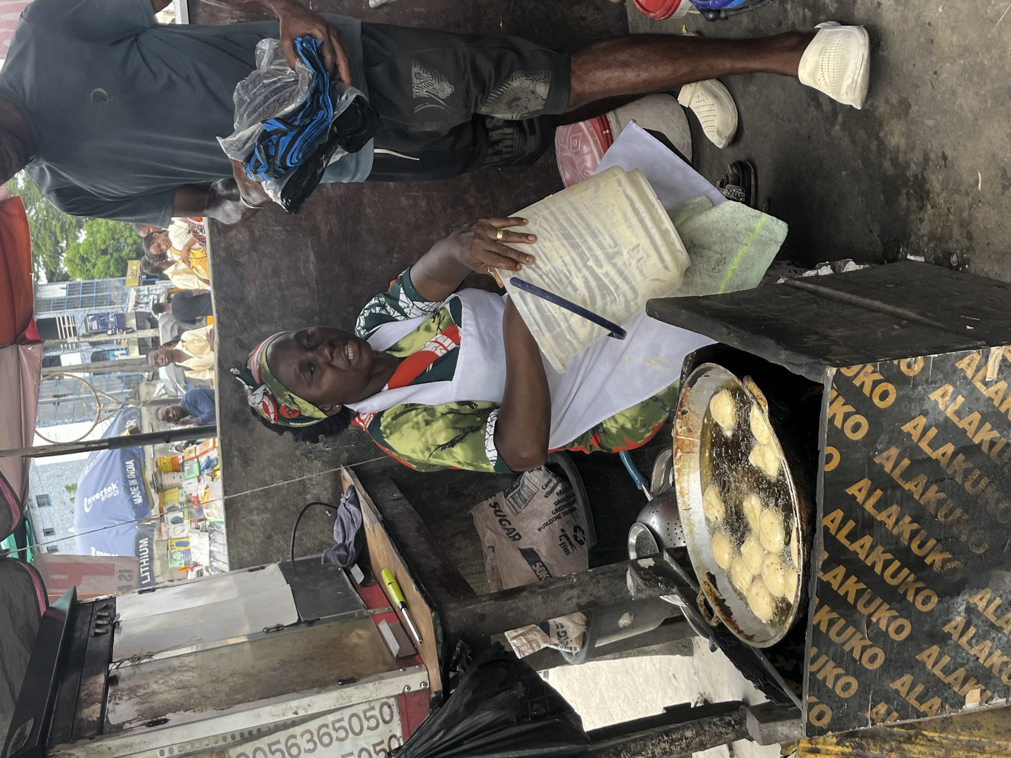 In Ado, Nigeria, a woman is cooking in a street food stall. She wears a headscarf, a green and white patterned top, and a white apron. She's smiling and looking upwards while holding a white container. A large frying pan filled with oil and round, doughy items sits on a burner. The stall is adorned with black and yellow signage bearing the repeated word "ALAKUKO." A bag of "SUGAR" is visible near her. A man stands beside her, holding a bundle of blue and black items. The surrounding environment includes other stalls, people, and buildings under an overcast sky.