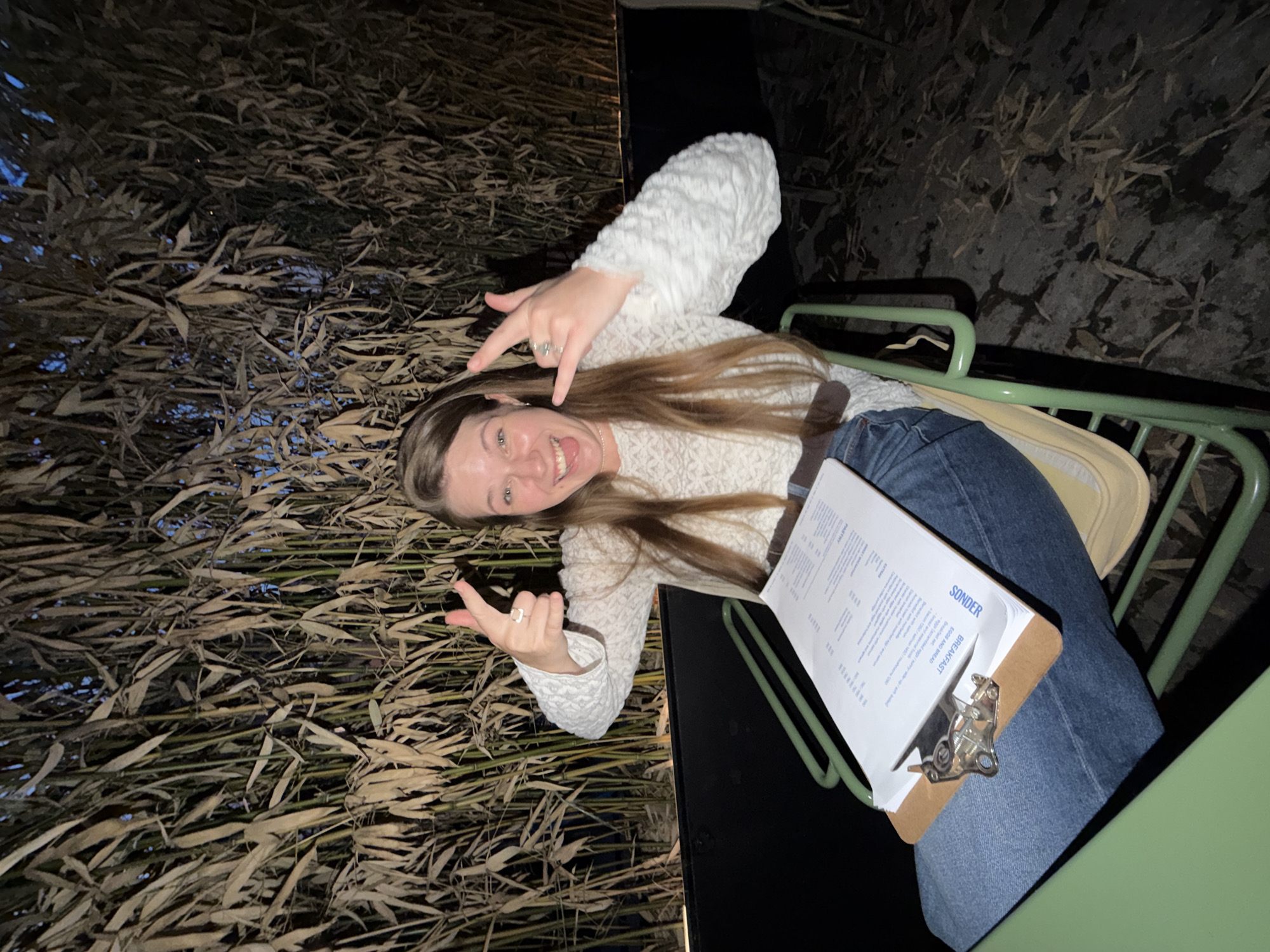 The image shows a woman sitting in a green metal chair at an outdoor restaurant in Belgrade, Serbia. She is holding a menu from a place called "Sonder". The area appears to be decorated with dried bamboo stalks. The woman is wearing a white fuzzy sweater and jeans. She is smiling and making a "rock on" hand gesture with both hands.