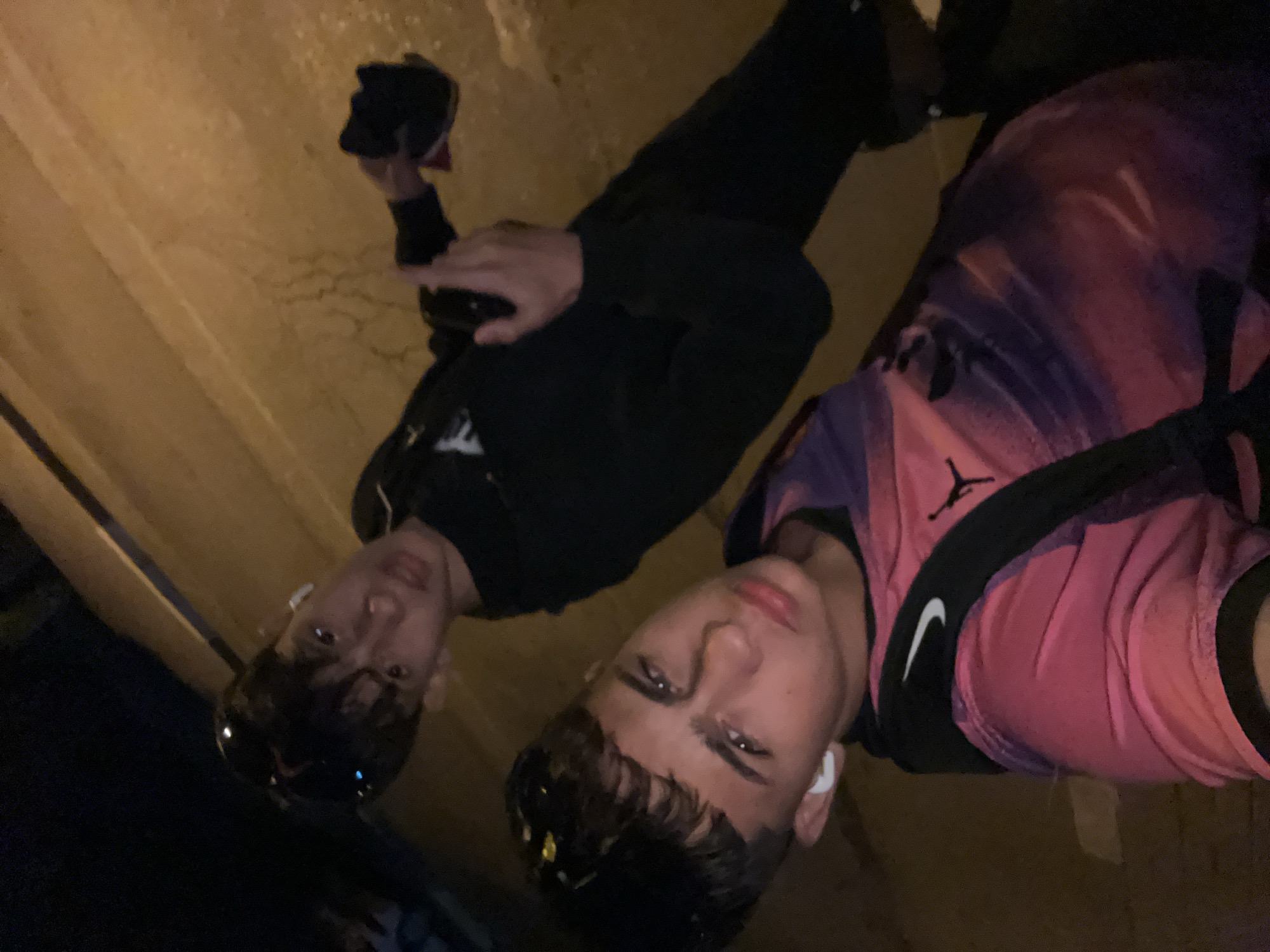 Two young males are captured in a low-light, outdoor setting, seemingly taking a selfie. The person in the foreground, closer to the camera, is wearing a pink and purple tie-dye patterned shirt with a black Nike swoosh logo visible on the shoulder. They have short, dark hair and are wearing white earbuds. Their gaze is directed towards the camera. Behind them, slightly out of focus, is another young male wearing a black hoodie. This individual is holding a dark-colored object in their right hand, possibly a phone. The background is indistinct but appears to be an outdoor urban environment, possibly a sidewalk or street, with a yellowish, textured surface suggesting pavement or a wall. The lighting suggests it is nighttime or a dimly lit area.