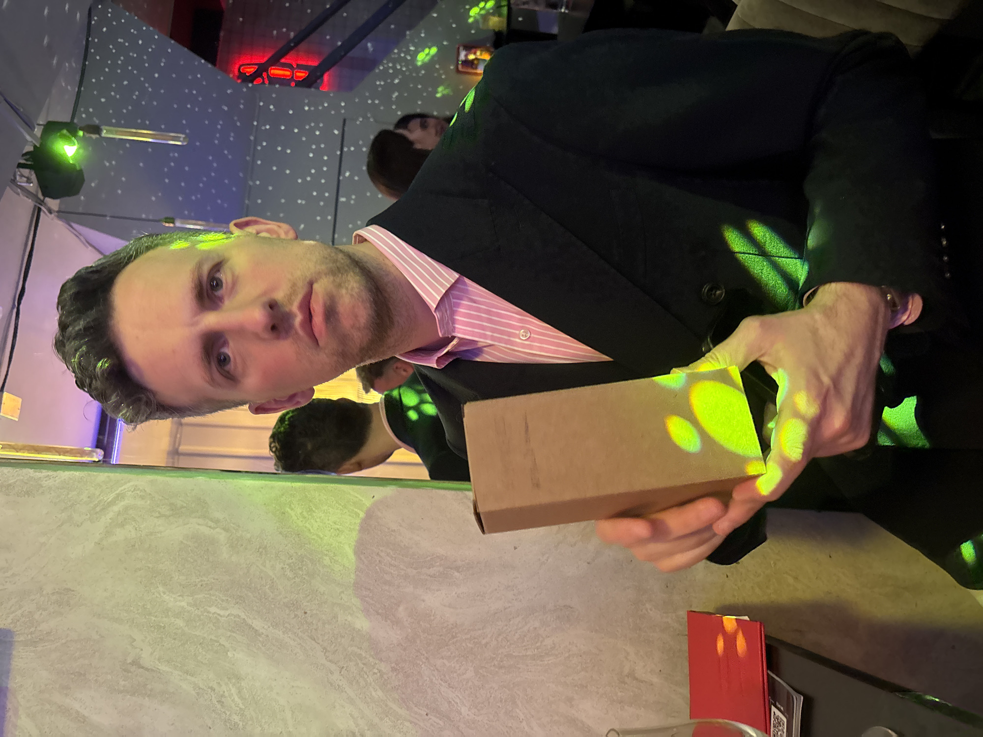 A man in a suit, wearing a pink and white striped shirt, is shown from the mid-chest up, holding a small cardboard box in his hands. Green and yellow lights are shining on his hands and the box. The background is a room with a mirrored wall. Reflected in the mirror is part of a table and the man’s face. Other people are in the background, but they are out of focus. There is a red object and some papers on the table. The ceiling has a pattern of small white lights, and there are other red and green lights visible. The location is in London, United Kingdom.