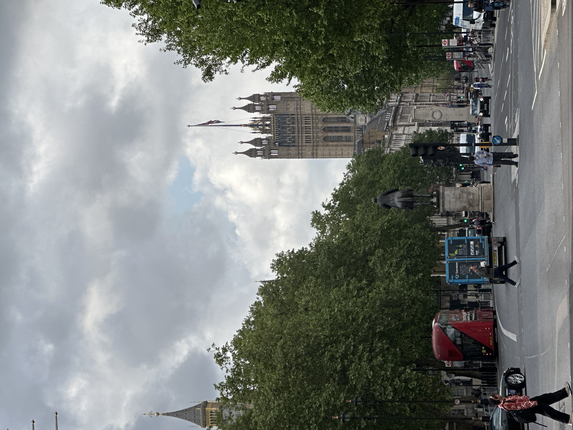 The image shows the Houses of Parliament in London, United Kingdom, partially obscured by trees. The Elizabeth Tower (Big Ben) is visible on the left. A British flag flies atop the Victoria Tower. A street runs along the right side of the frame, featuring a red double-decker bus, cars, street signs, and pedestrians. The sky is overcast with clouds. A statue is visible in the foreground near the trees. People are crossing the street.