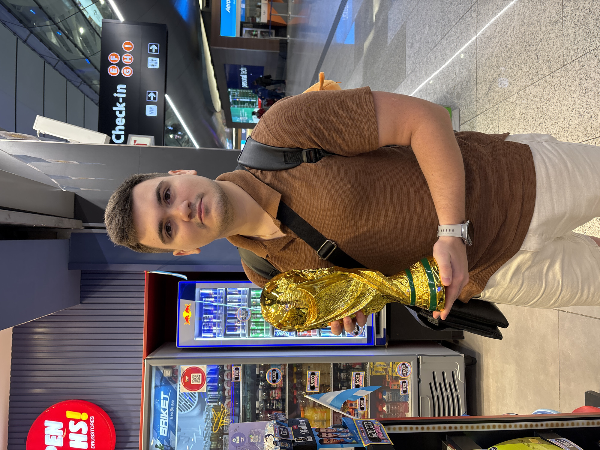 A young man stands in an airport terminal, holding a replica of the FIFA World Cup trophy. He is wearing a brown polo shirt and light-colored shorts. A black backpack is slung over his left shoulder. To his left, there are two refrigerators, one stocked with various beverages and the other with Red Bull cans. An Argentinian flag is placed on a shelf in front of the refrigerators. A sign in the background reads "Check-in" with directional arrows and lettered counters. The floor is tiled, and in the distance, there are other travelers and airport signage. The man appears to be in a public area of Aeropuerto Internacional Ezeiza, Argentina.