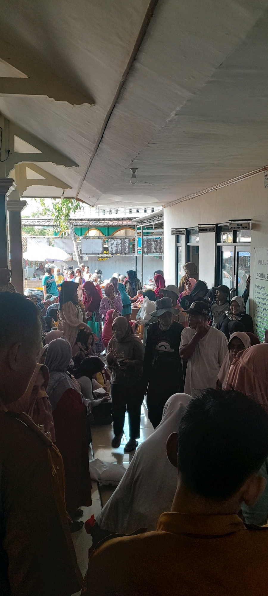 The image depicts a crowded indoor scene, possibly a community center or a government office, in Jeruklegi Kulon, Indonesia. A large group of people, predominantly women wearing hijabs, are gathered in a shaded, open-air area. The architecture suggests a tropical climate with an extended roofline supported by pillars. Natural light streams in from the left, illuminating a portion of the gathering. Some individuals are standing, while others appear to be seated or waiting. The background reveals more buildings with patterned facades and foliage, hinting at an outdoor space beyond. Signs with Indonesian text are visible on the building walls, including what appears to be a list of services or procedures. The overall atmosphere is one of a busy, public gathering.