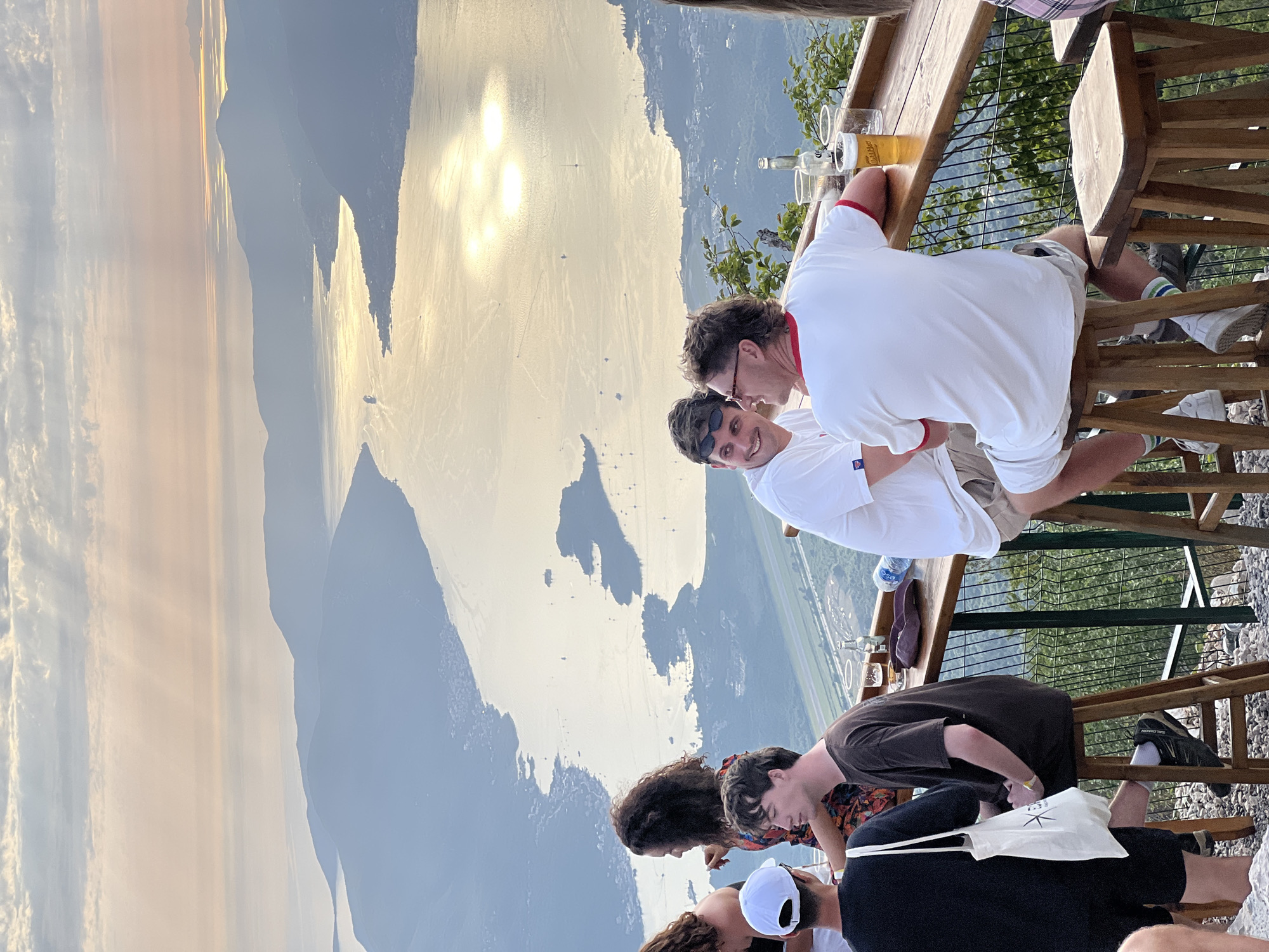 The scene is situated in the city of Kuk, Montenegro, capturing a panoramic view from an elevated outdoor bar at what appears to be sunset or golden hour. The sky exhibits soft clouds with distinct crepuscular rays, casting ethereal light across the landscape. In the midground, a large, winding bay or fjord dominates the view, its surface reflecting the intense golden sunlight in shimmering streaks and patches. Numerous small boats, including sailing vessels, are scattered across the water. The bay is framed by a series of steep, hazy blue-gray mountains and hills that recede into the distance, with small towns and settlements visible along the coastlines. A larger ship is discernible further down the bay. In the foreground, an outdoor bar area is set up with a long, light brown wooden counter and tall, square-seated wooden stools. A dark metal railing with a grid pattern separates the patrons from a steep, tree-covered slope descending into a valley below. The ground beneath the bar stools is rocky. Several individuals are gathered at the bar: - On the far left, a person wearing a black t-shirt and shorts, with a white baseball cap worn backward, is partially visible from the back, carrying a white canvas tote bag featuring a dark, spiky star logo. - Next to them, a woman with voluminous dark curly hair wears a colorful patterned dress and is seen looking towards a man seated beside her, her right arm slightly raised in an apparent gesture. - The seated man, with short dark hair, wears an oversized dark brown t-shirt, dark trousers, and black Salomon sneakers with white socks. He is perched on a wooden stool, facing partially away but engaged with the woman. An empty clear glass rests on the bar in front of him. - To the right, another man with short dark hair, wearing a white t-shirt and light shorts, has sunglasses pushed onto his head. He is smiling and looking towards the man further right, engaged in conversation. - The man on the far right has a prominent mullet hairstyle, wearing a white t-shirt with distinctive red trim on the collar and sleeves, light-colored shorts, and white sneakers with green and white striped socks. He also has sunglasses on his head and is actively conversing with the man to his left, with a relaxed expression. In front of him on the bar are a glass of