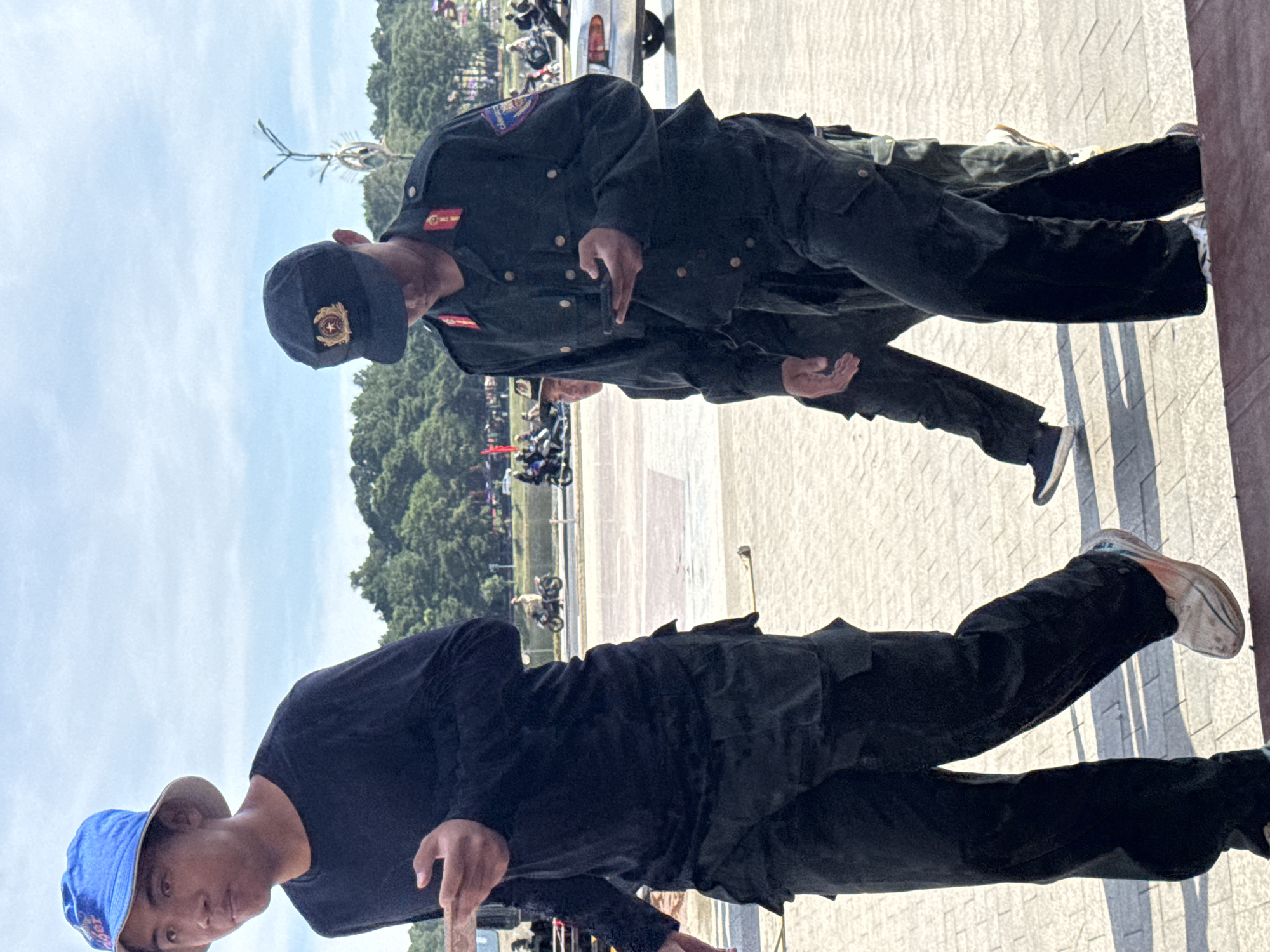 A group of individuals, including two in police uniforms, are walking up a paved walkway under a cloudy sky. The individual in the foreground on the left wears a black long-sleeved shirt, black cargo pants, and a blue bucket hat. They are holding a smartphone in their left hand. To their right, a police officer in uniform is also looking at a smartphone. They wear a dark uniform with red and gold insignia on the collar and a dark cap with a red and gold emblem. Behind them, another person in a similar dark uniform is visible. The walkway is made of light-colored rectangular paving stones. In the background, there are trees, streetlights, vehicles, and other people. The scene appears to be in a public area, possibly a park or plaza, in Da Lat, Vietnam, suggested by the presence of Vietnamese police uniforms and the tropical foliage.