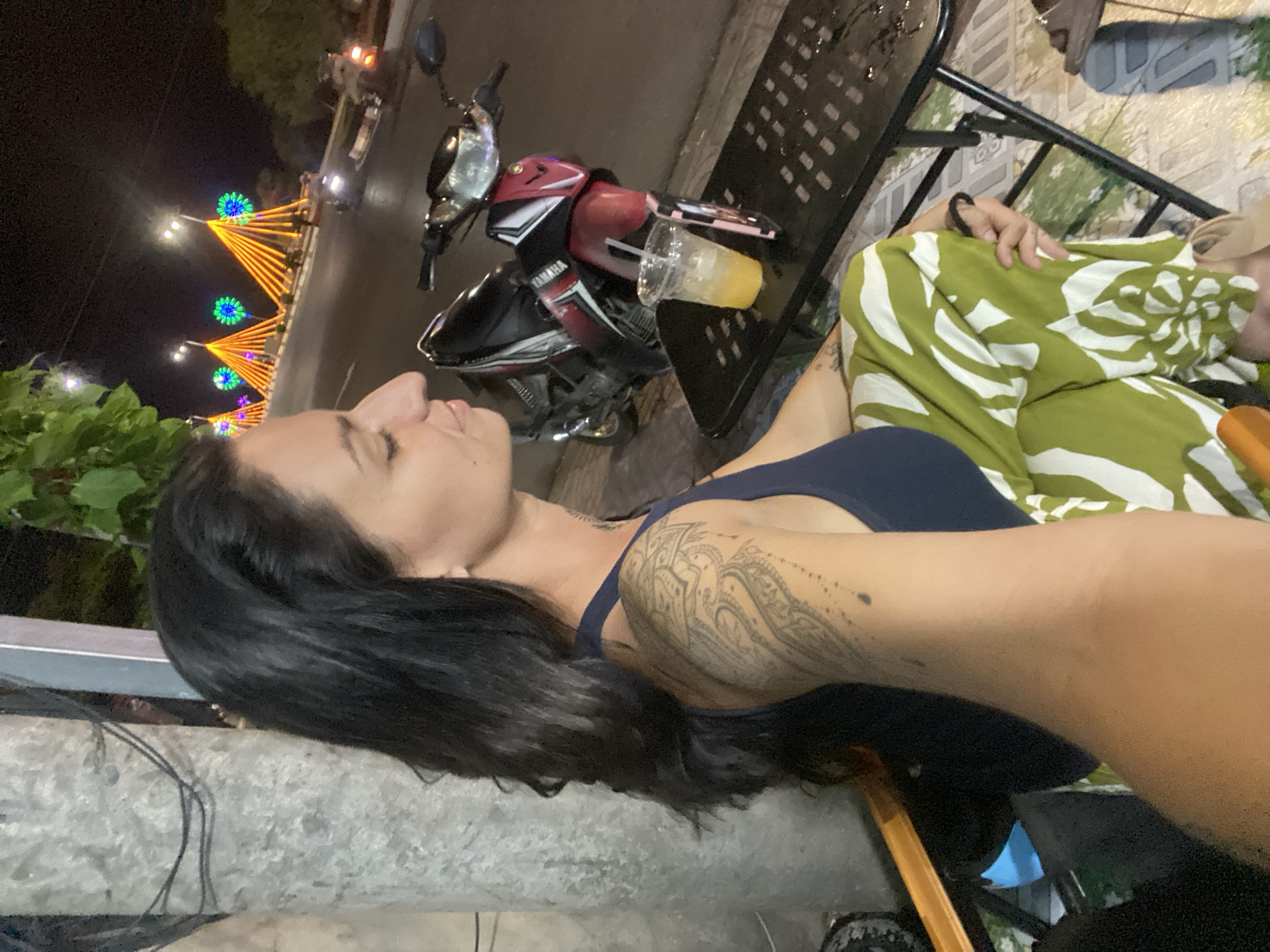 A medium shot captures a woman with long, dark hair sitting at a table in Phu Thuy Ward, Vietnam, at night. She is wearing a dark blue tank top and green and white patterned pants. Her left arm, adorned with a large tattoo, rests on the table. In front of her is a clear plastic cup filled with an orange beverage and ice, and a cell phone. A black metal table and chairs are visible. Behind her, a red and black Yamaha motorcycle is parked. The background features a street illuminated by decorative orange and green lights, suggesting a festive atmosphere. The overall scene is an outdoor setting, likely a cafe or street-side eatery.