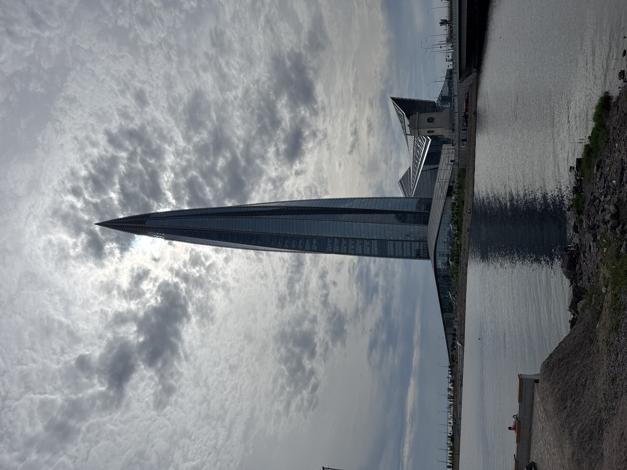 The image captures a view of the Lakhta Center in Saint Petersburg, Russia, set against a sky filled with textured clouds. The skyscraper's sharp, triangular peak pierces the cloudy sky, and the building's facade reflects the ambient light. To the right of the Lakhta Center, there is a body of water with a rocky shoreline. Along the water's edge, there is a promenade with streetlights. A small portion of a paved walkway with a bench is visible at the bottom right corner of the image, where a person in pink clothing is sitting on the bench.