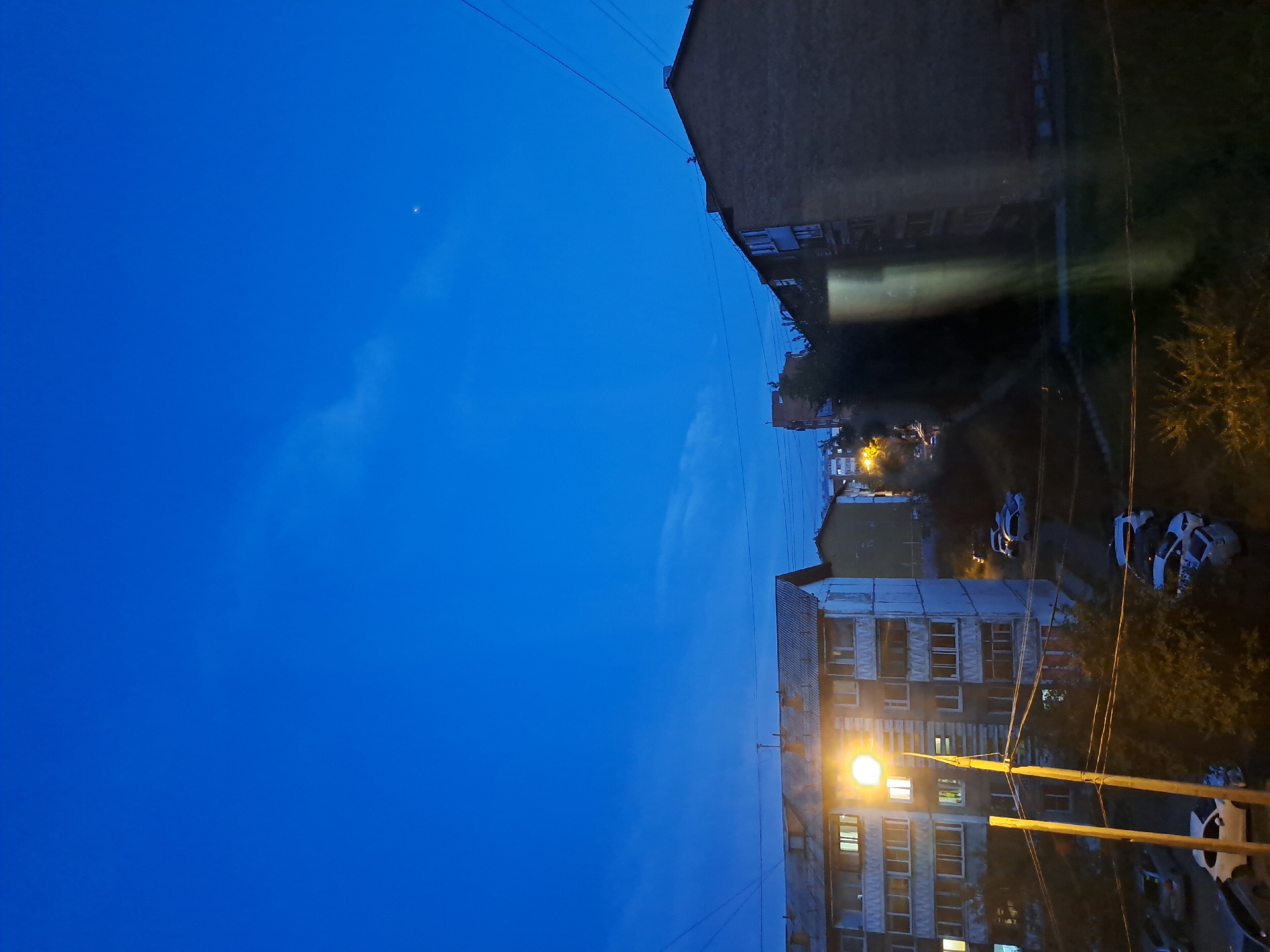 The image captures a twilight scene in an urban setting. In the foreground, a street lamp illuminates a residential building with multiple windows and balconies, casting a warm glow. Several cars are parked in front of the building and on a path winding through the scene. In the background, other buildings and trees are visible, silhouetted against the deep blue sky. A faint light, possibly a star or planet, is visible in the upper right portion of the sky. The overall impression is one of a quiet evening in a city neighborhood.