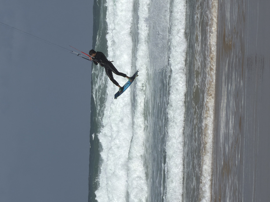 A male individual in a black wetsuit is captured mid-air while kitesurfing over the Mediterranean Sea, off the coast of Tel-Aviv, Israel. The subject leans backward and to the right, with both feet secured to a teal and black kitesurfing board. His arms are extended forward, gripping a dark control bar with red accents on its right side, from which multiple lines ascend out of the frame, connected to an overhead kite. Below the airborne kitesurfer, the turbulent grey-green water is marked by numerous breaking, white-capped waves extending into the distance. A strip of wet, dark sand is visible at the bottom of the frame, indicating the immediate shoreline. The sky above is uniformly overcast and dark grey.