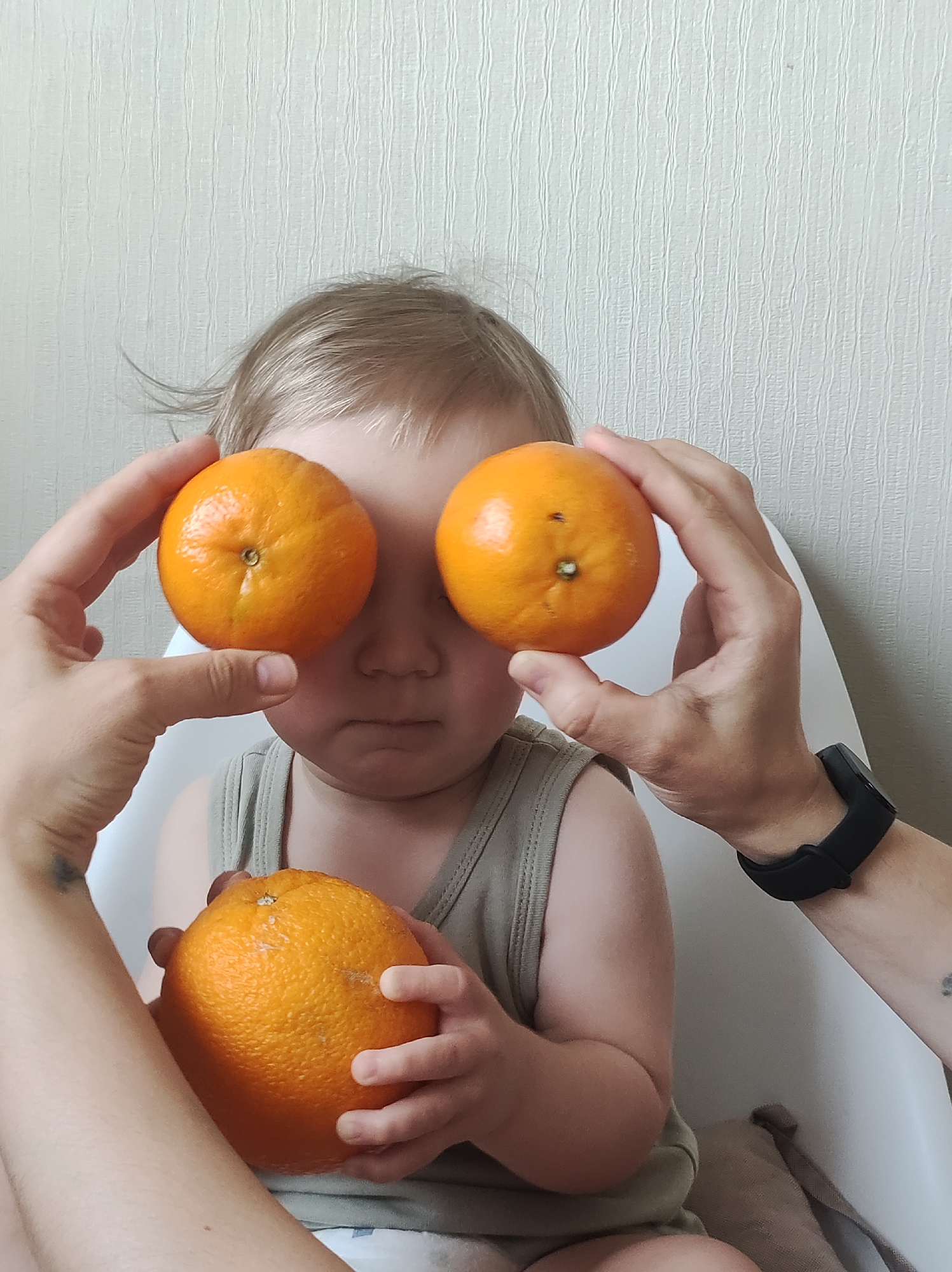 A close-up shot shows a young child sitting in a white high chair. The child has fair hair and a light-colored tank top. Two hands hold oranges up to the child's eyes, effectively covering them. Another orange is held by the child in their lap. The background is a textured, off-white wall. The scene suggests a playful moment between the child and an adult. The location is Unknown, Unknown.
