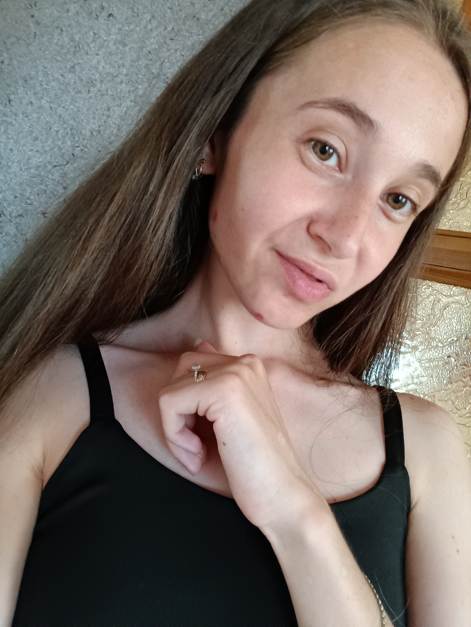 This is a close-up, self-taken photograph of a young woman with long, brown hair and light brown eyes. She is wearing a black tank top and has her left hand resting near her chin, adorned with two rings. A small red mark is visible on her chin, and another small mark is visible on her neck. The background appears to be an interior setting with a textured wall and a patterned surface to the right. The photograph was taken in Vypasne, Ukraine.