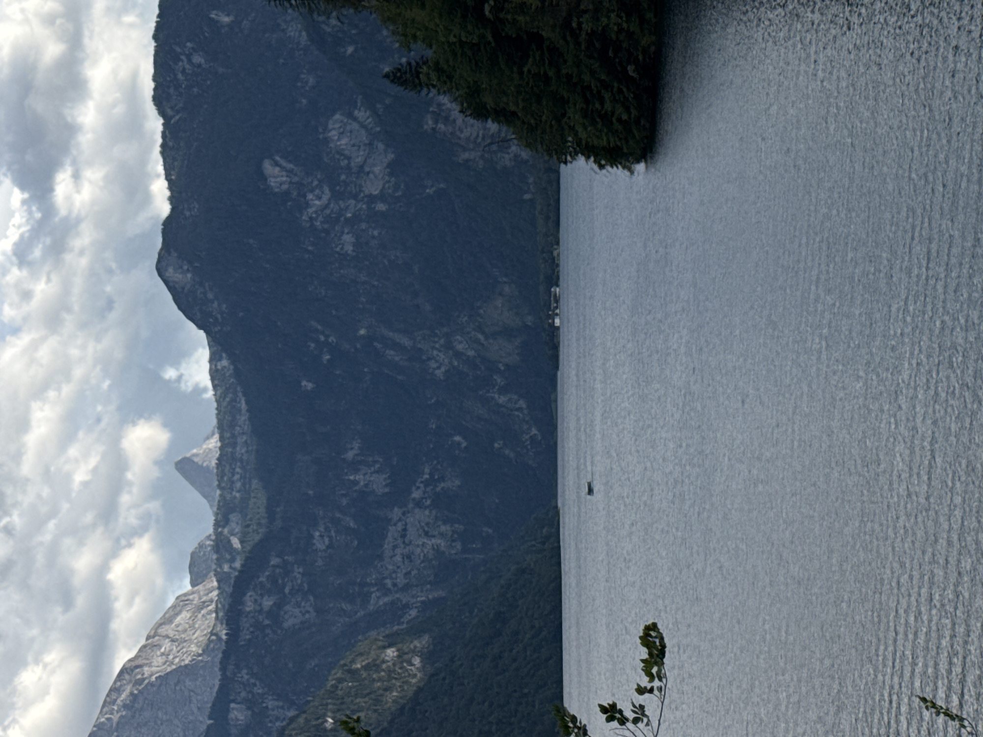The image shows a serene view of Königssee lake in Schönau am Königssee, Germany. The calm, grey-blue water of the lake fills the foreground and midground. In the distance, a small white building or house is visible on the shore, nestled amongst dark green trees that line the edge of the lake. Towering, dark green, forested mountains rise steeply from the opposite shore, their rugged slopes partially obscured by shadows. Above the mountains, a dramatic sky with heavy, grey clouds is visible, suggesting overcast weather or the approach of a storm. A single, small, dark boat is positioned on the water, adding a touch of human presence to the vast natural landscape. The overall scene is one of tranquil, imposing natural beauty.