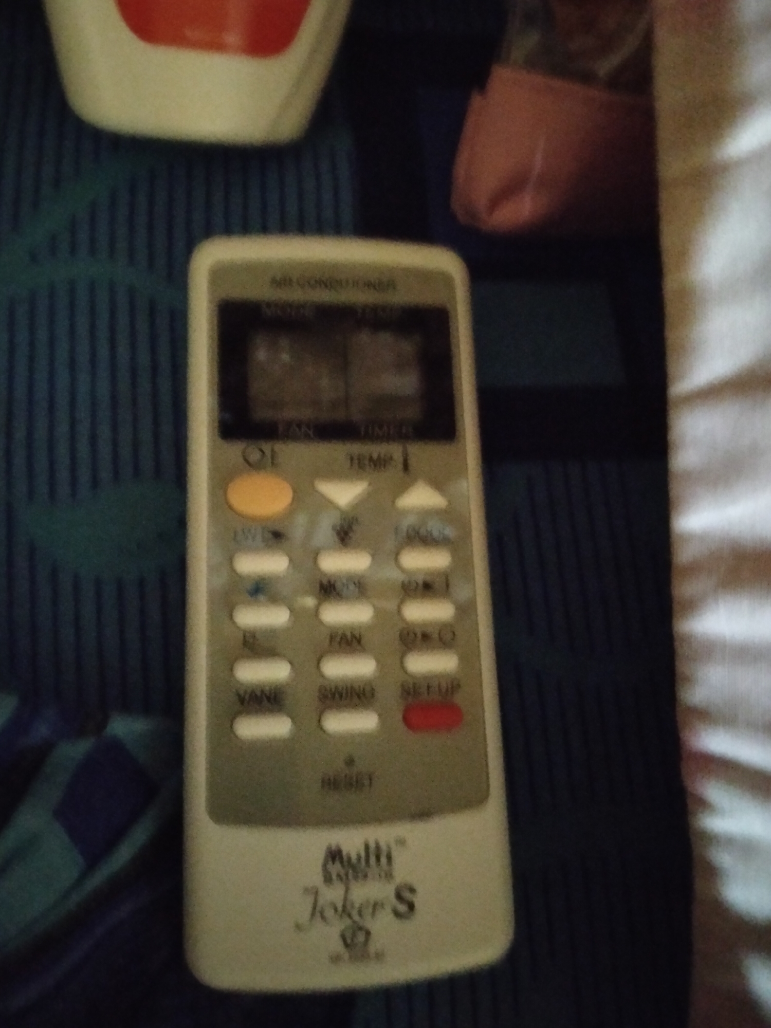 An overhead shot shows a white air conditioner remote control with a digital display and various buttons. The remote is positioned on a dark blue textured surface. To the left of the remote is a hint of a blue and green striped fabric. To the right of the remote, a white textured surface is visible. The remote control has the brand name "Multi" printed on it, followed by "Joker S". Buttons on the remote include "TEMP", "MODE", "FAN", "VANE", "SWING", and "RESET". The digital display shows two numerical values, one above the other. The image is undated and unlocated in Payagaboh, Indonesia.