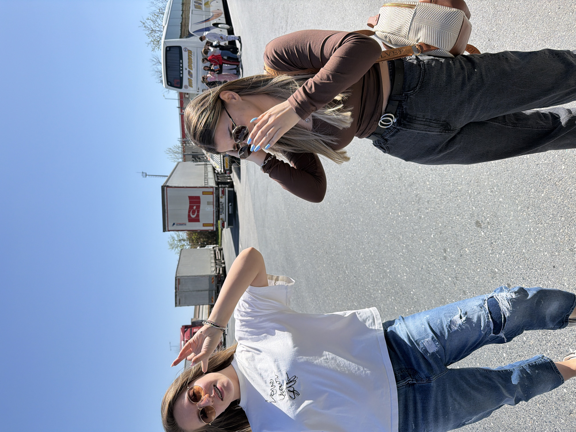 The image depicts two young women in an outdoor setting. The woman on the right is wearing a brown long-sleeved top, dark gray jeans with a unique double-circle belt buckle, and a backpack. She is putting on sunglasses and has blue nail polish. The woman on the left is wearing a white t-shirt with an embroidered design and ripped blue jeans. She is wearing sunglasses with round lenses and making a peace sign. In the background, there is a bus filled with people and several trucks, one of which has a Turkish flag on its side. The setting appears to be a parking lot or rest area near a roadway. The scene's location is Çekmeköy, Turkey.
