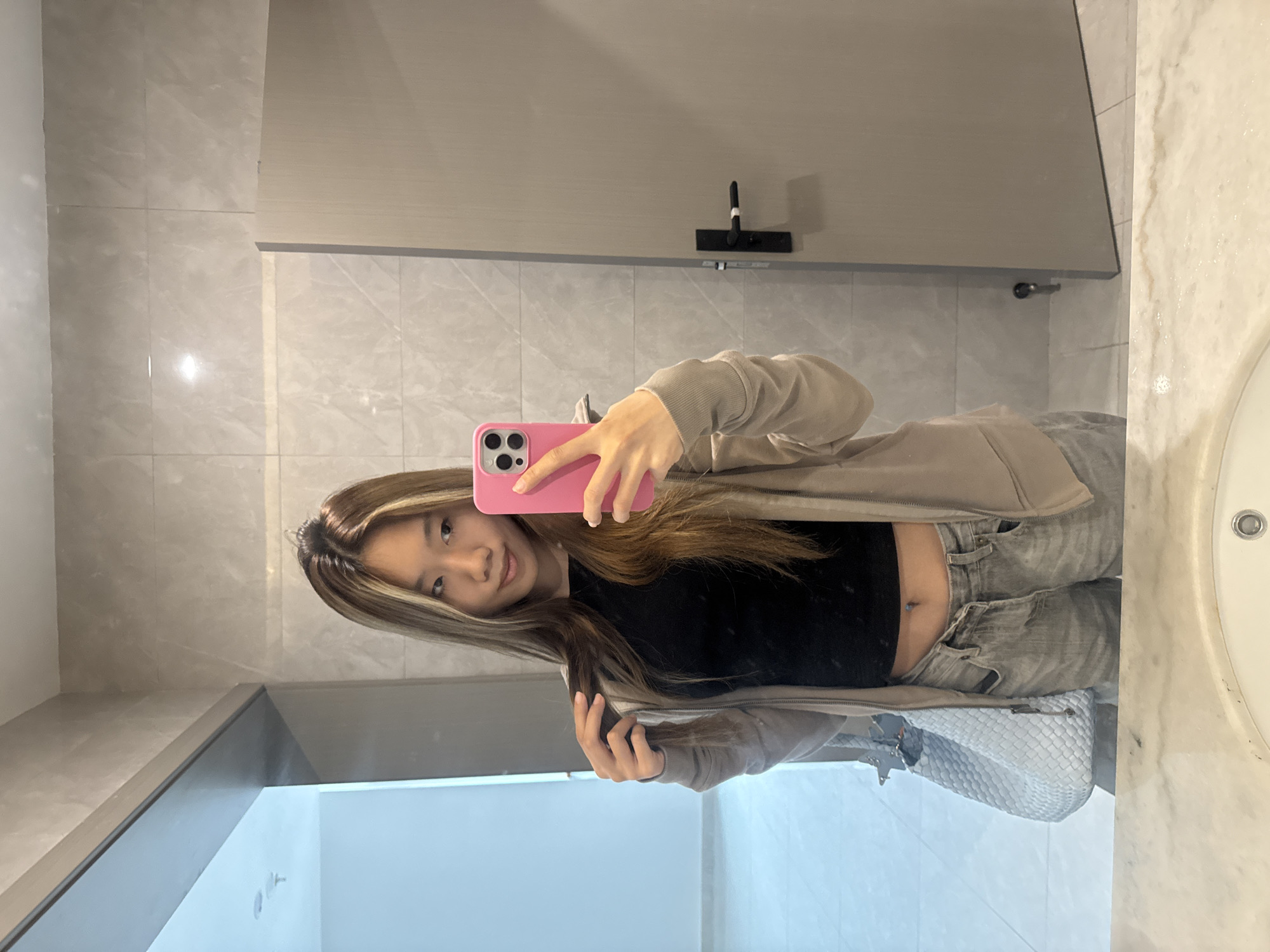 A young East Asian woman is featured in a mirror selfie, captured within what appears to be a public restroom in Nan'an District, China. She possesses long, straight brown hair with prominent blonde highlights. She is dressed in a black crop top, revealing her midriff where a small, light blue belly button piercing is visible. Over the crop top, she wears a light beige or tan zipped hoodie, paired with light-colored, possibly grey, pants. She holds a pink smartphone, likely an iPhone, in her right hand to take the photo, while her left hand gently touches her hair near her shoulder.
The setting consists of light grey marble-patterned wall tiles and large, plain light grey panels, possibly indicating restroom stalls or architectural features. A portion of a white sink basin is visible on the right side of the frame. In the background, mounted on a wall and reflected in the mirror, is a black T-shaped object with a white tip, potentially a hand dryer or dispenser. A light-colored, possibly white, woven handbag with a metallic clasp is visible positioned below her waist. The woman exhibits a soft, closed-mouth smile, suggesting a relaxed and pleasant mood. The lighting is artificial, typical of an indoor public space, and no specific time of day or weather conditions can be discerned. There is no visible text within the image.