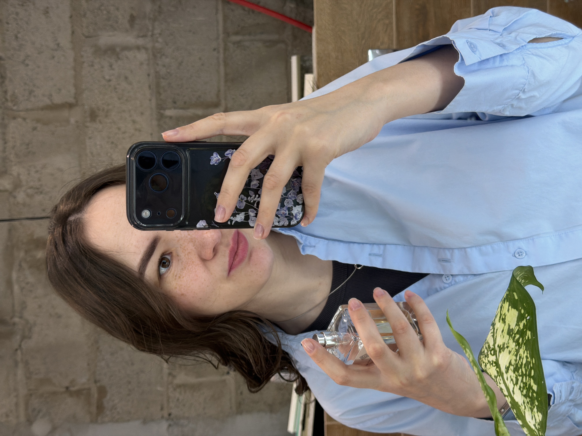 A young woman with light skin and freckles is taking a selfie using a black smartphone with a floral-patterned case. She is wearing a light blue button-down shirt and a black top underneath. She holds a clear glass perfume bottle in her right hand. The background appears to be an interior with a brick wall and possibly some furniture or objects partially visible. A green and white plant is also visible next to her. The location is unknown and cannot be identified as the city of Chaikovsky, Russia based on the image.