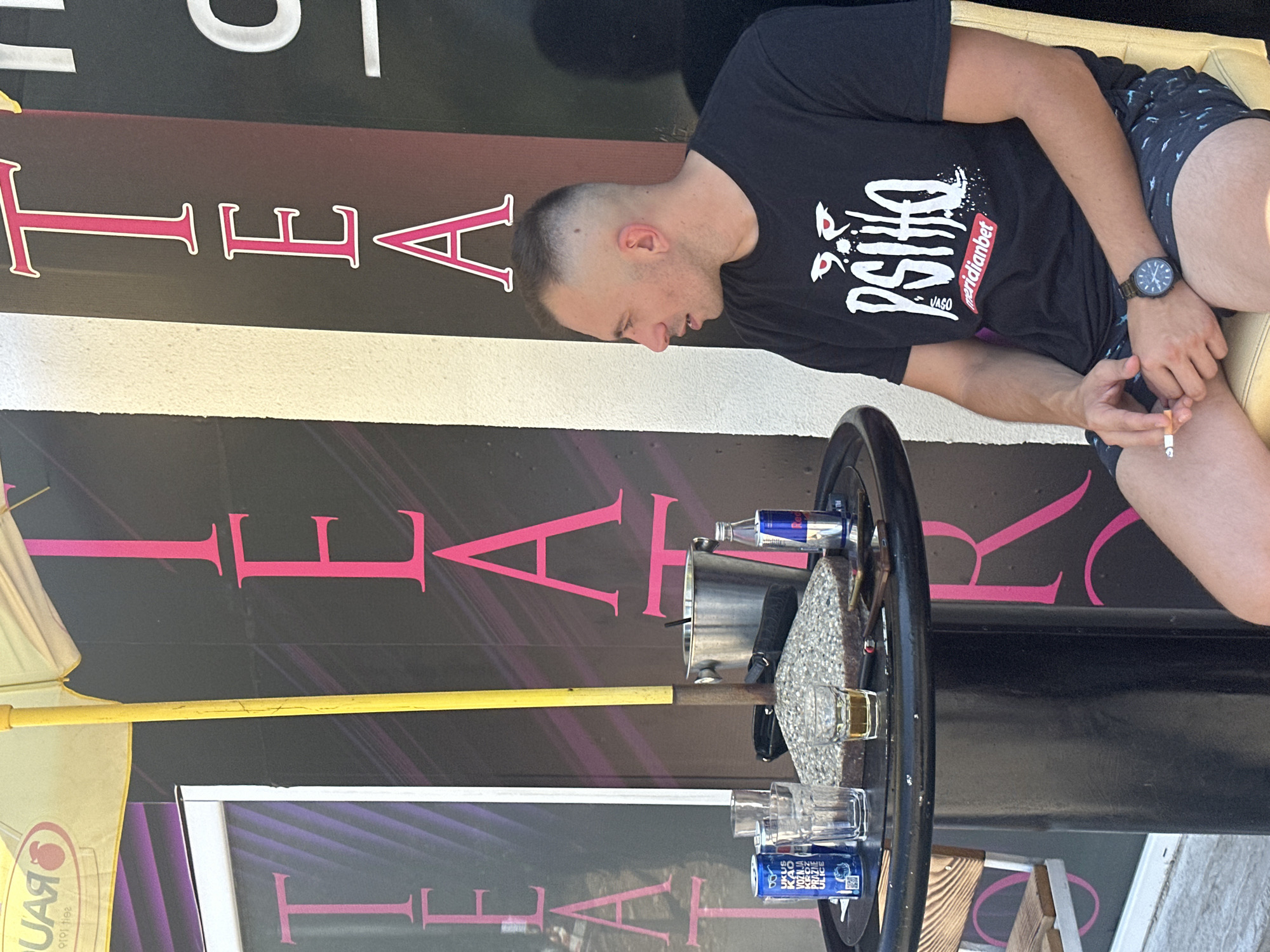A man is sitting at a round black table with drinks and a cigarette. He is wearing a black t-shirt with white writing and a digital watch on his left wrist. The man is holding a cigarette in his right hand. On the table there are three glasses, a can of blue drink, a red bull can, a silver bucket with ice, and a small bottle of alcohol. In the background, there is a wall with large pink letters that spell "TEATRO", and a yellow umbrella. This scene is taking place in front of a building on the street in the city of Budva, Montenegro.