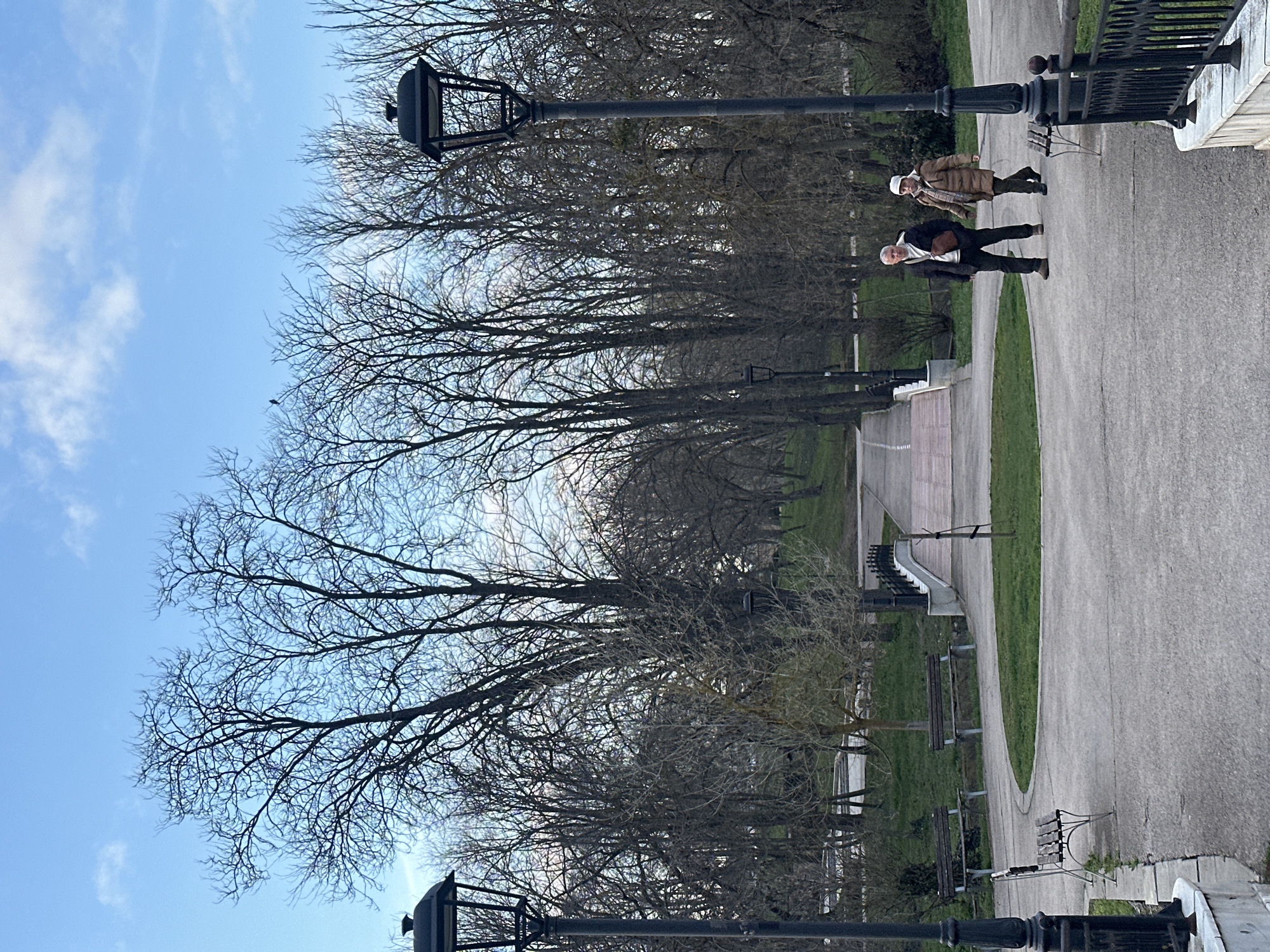 The image depicts a park scene in Dobrich, Bulgaria. Two people are walking on a paved path lined with bare trees and lampposts. Benches are spaced along the path. The sky is light blue with scattered clouds. A bird is perched on a tree branch.