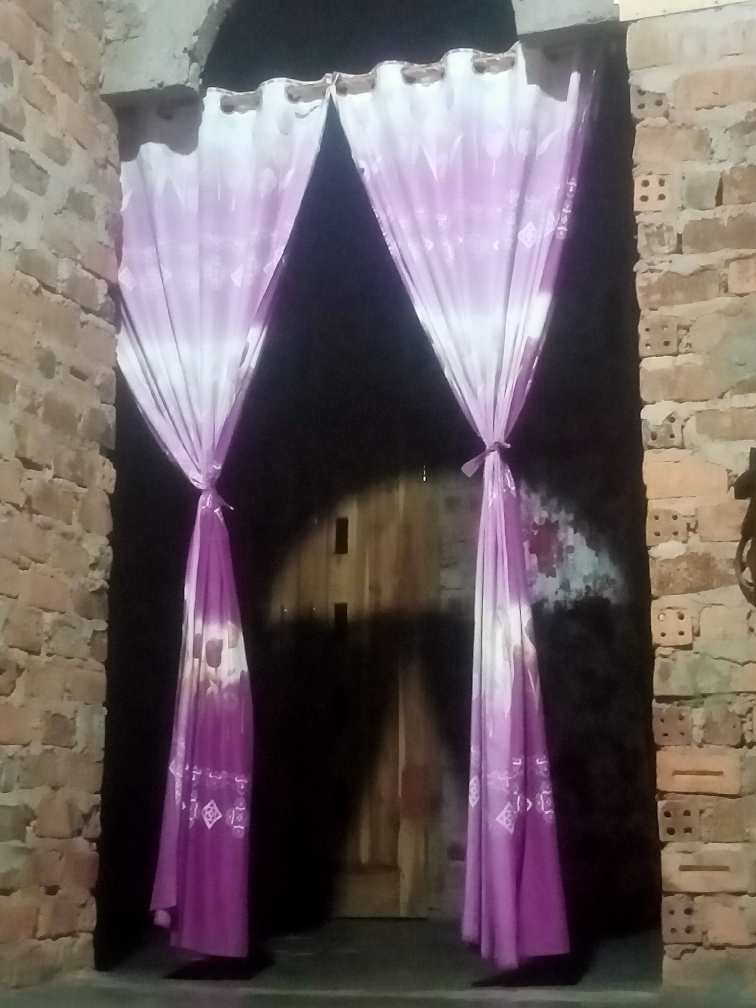 The image shows a doorway framed by brick walls, with two draped curtains made of light purple and white fabric. The curtains are tied back with fabric ties, creating a V-shape. A wooden door is visible in the background. The ground in front of the doorway appears to be a concrete or paved surface. The lighting is dim, suggesting it might be evening or night. The location is identified as Nusa Agung, Indonesia.