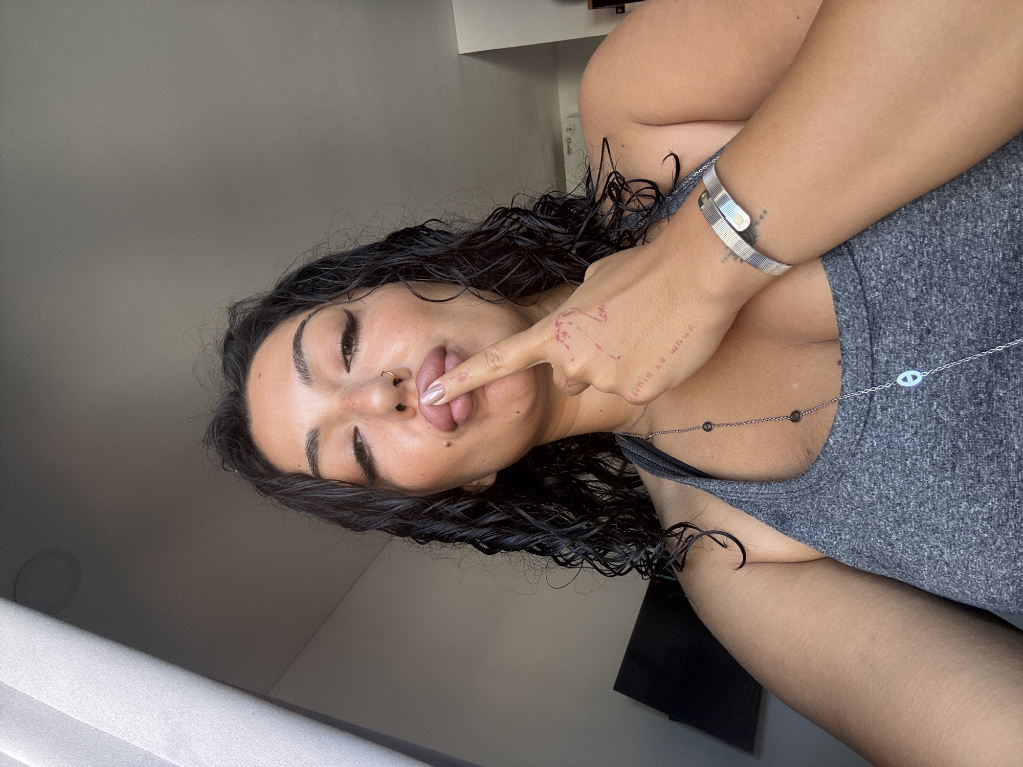 The image shows a close-up of a woman's face and upper body. She has dark, curly hair and is wearing a grey tank top. Her finger is touching her lips. She is wearing a silver bracelet and a necklace with a peace sign pendant. The background is out of focus, but appears to be an indoor setting with a window to the left. There are no discernible elements in the image that would suggest a specific location in Kuta Selatan, Indonesia.