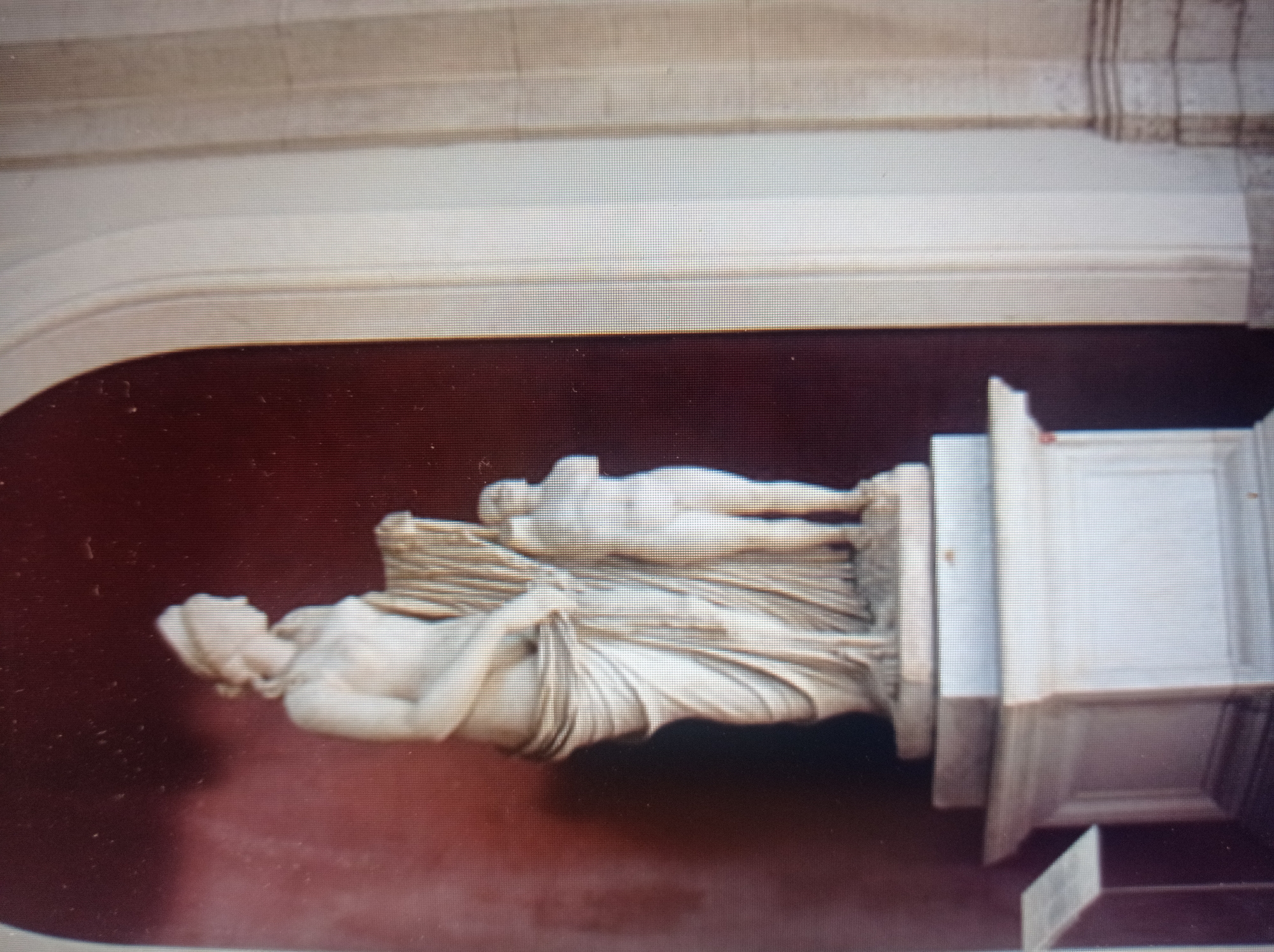 The image shows a marble statue of a woman and a child, likely mythological figures, displayed within an architectural niche. The statue is positioned on a multi-tiered white pedestal. The background wall is a deep red, and the right side of the frame features architectural elements like a fluted column and an arched opening. The lighting is somewhat dim, highlighting the textures of the marble and the drapery of the statues. There is a small information placard to the left of the pedestal. The style of the statue suggests classical art. The location is not specified as Mukachevo, Ukraine.