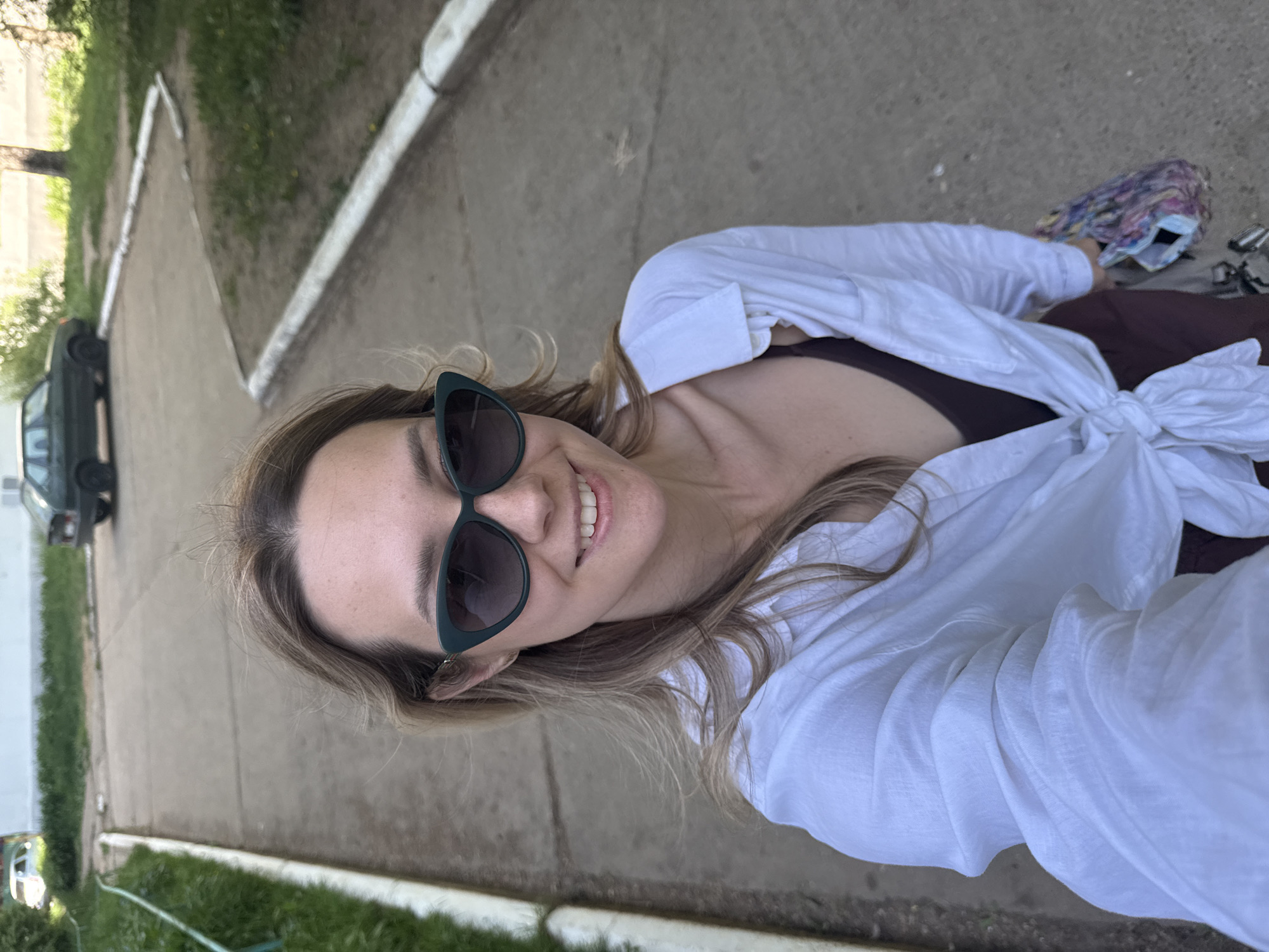 A fair-skinned woman smiles while taking a selfie outdoors on a concrete sidewalk. She is wearing dark sunglasses and a white button-down shirt knotted at the waist over a dark top. In the background are grassy areas and trees, a road, a parked car, and curbs. The photo was taken in Chaikovsky, Russia.