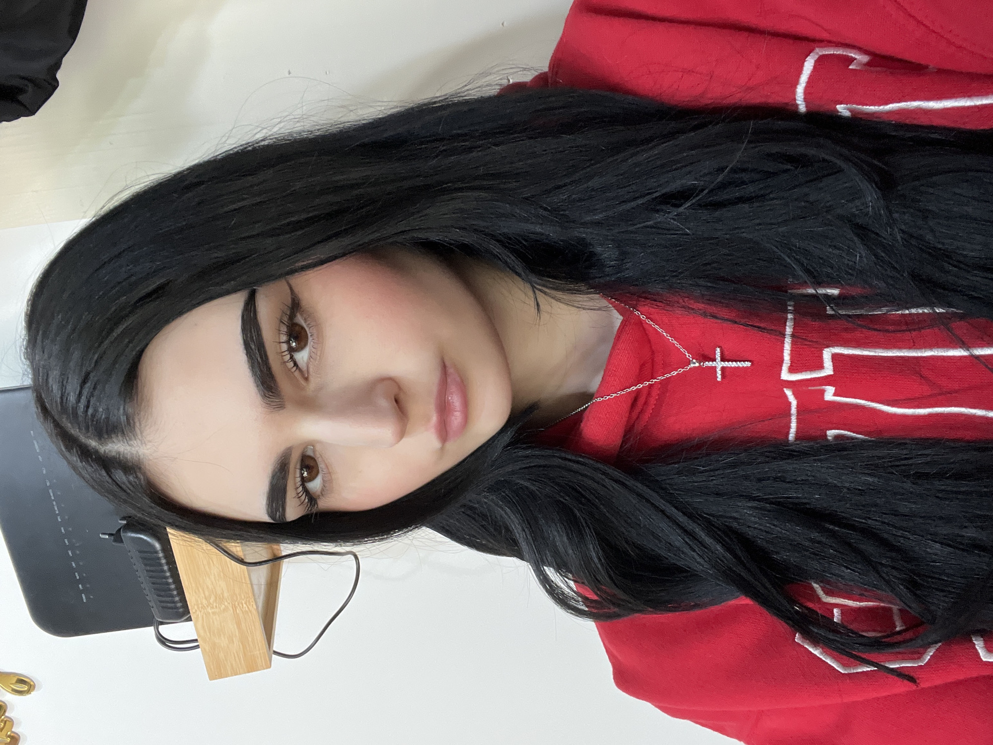 A woman with long, black hair and light skin is facing the camera, tilted slightly to her right. She has dark eyebrows and brown eyes. She is wearing a red hooded sweatshirt with white lettering. A silver necklace with a cross pendant is visible around her neck. In the lower left corner of the image, there is a black electronic device with a wooden stand and a black cord attached. The background is a plain white wall. The location is not discernible from the given image and cannot be determined to be in Burgas, Bulgaria.
