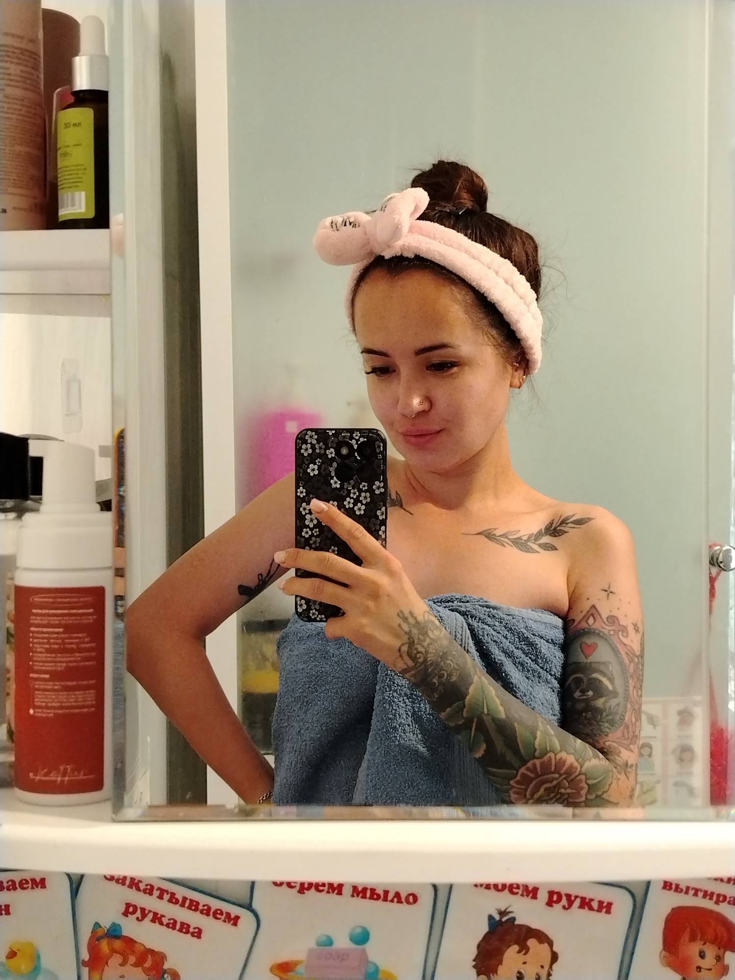 The image shows a young woman with dark hair pulled up in a bun, wearing a pink headband and wrapped in a blue towel. She is taking a selfie in a mirror, holding a black phone with a floral pattern. She has tattoos on her arms and shoulder. The background appears to be a bathroom with shelves containing bottles and other toiletries. There are also decorative cards with illustrations of children and text in Russian visible on a surface below the mirror. The location in the city of Noviy, Russia is not determinable from the image.