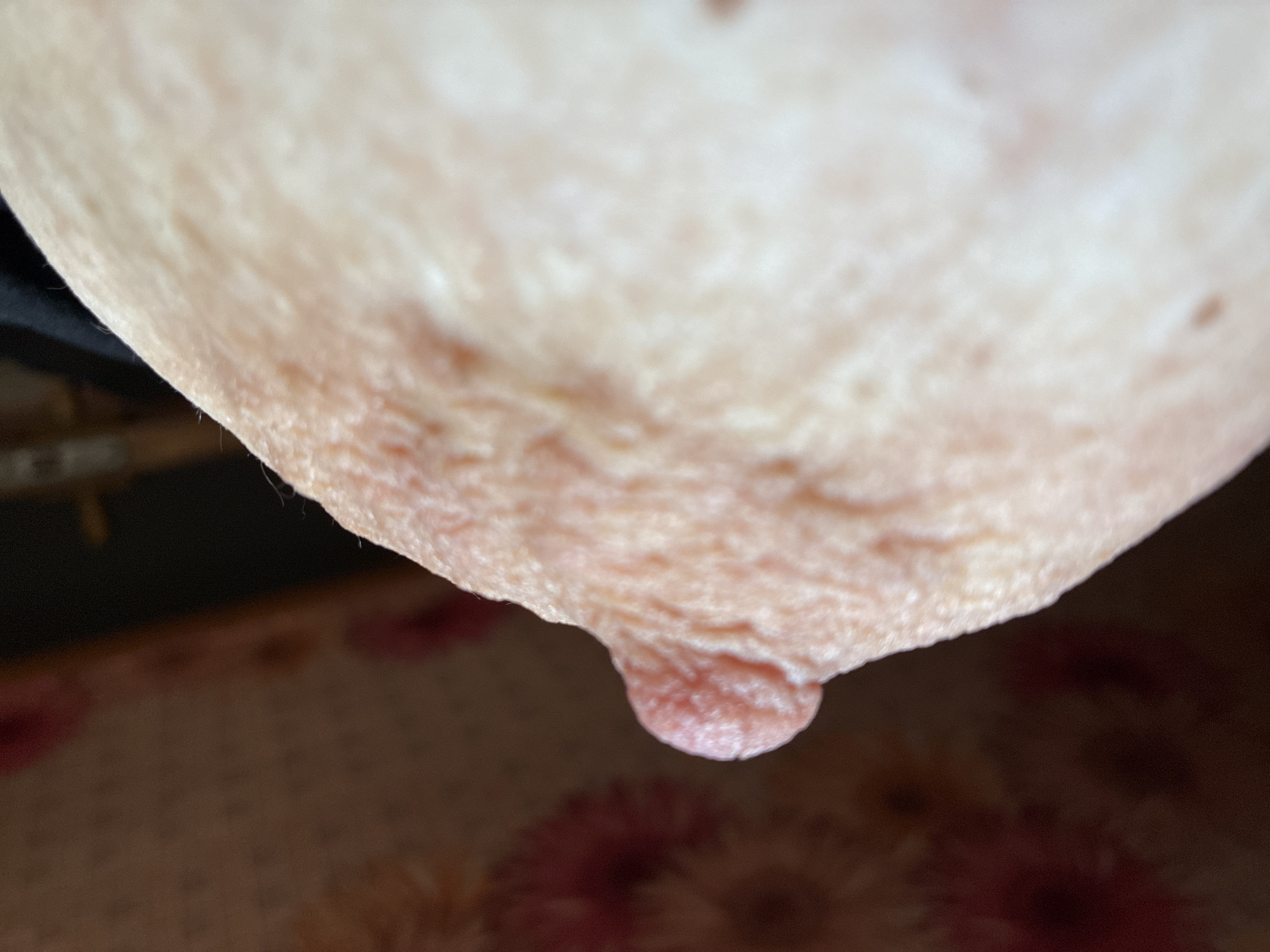The image is a close-up, extreme macro photograph of a human nipple and areola, specifically showing the texture of the skin and the nipple protrusion. The background is out of focus, with a patterned fabric on the left and a dark area on the right. There are no discernible geographical markers or indications of the city of Zhirnov, Russia, within the image.