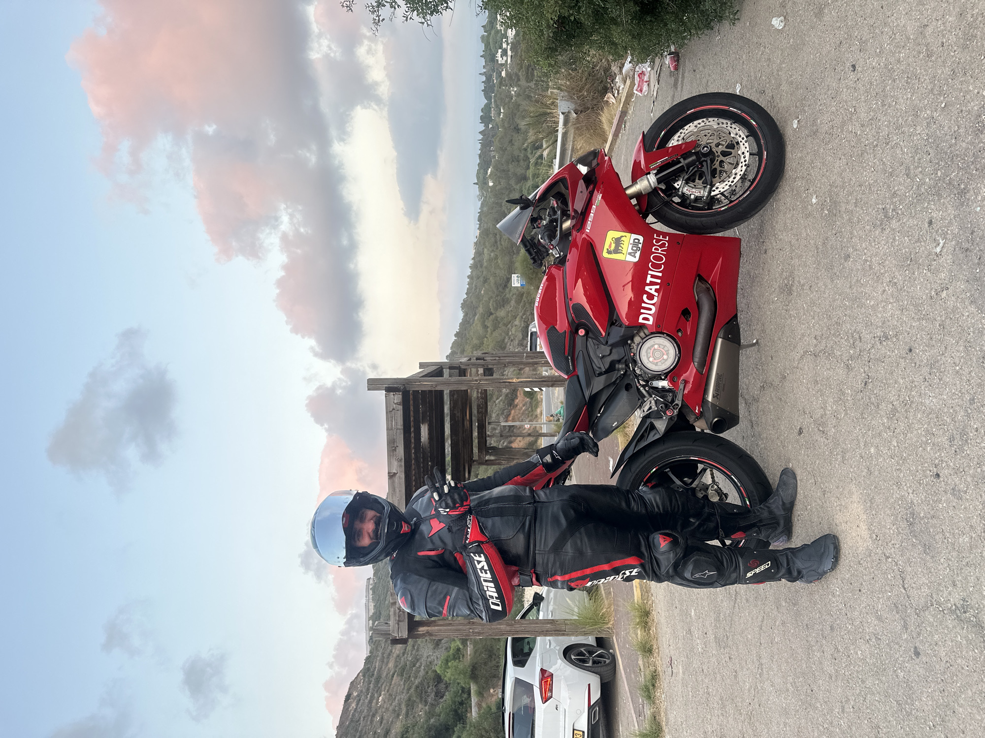 A person wearing a full black and red leather motorcycle suit, including a helmet with a mirrored visor, stands next to a red Ducati motorcycle. The person is smiling and making a peace sign with their hand. The motorcycle has "DUCATI Corse" branding on the side and an "Agip" sticker. In the background, there is a white car, a wooden structure, a hillside covered in trees and vegetation, and a distant view of buildings. The sky is partly cloudy with hints of pink and orange from what appears to be sunset. The scene is set on a paved area next to a road, with some litter visible on the ground. The location is identified as Mate Asher Regional Council, Israel.