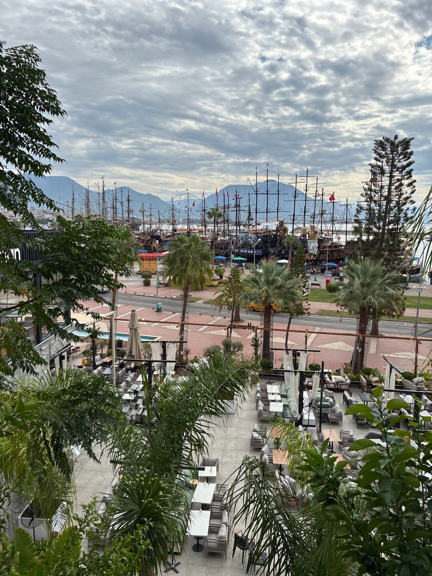 The image captures a panoramic view of the Alanya Marina in Alanya, Turkey, on a cloudy day. In the foreground, lush green palm trees and other foliage partially obscure a modern outdoor seating area with white tables and gray upholstered chairs arranged on a tiled patio. Several white gazebos with draped fabric provide shade over some of the seating arrangements.
Beyond the patio, a street runs parallel to the waterfront, with palm trees lining the roadside. On the other side of the street, a brick-paved walkway is visible, dotted with more palm trees and streetlights. Further back, a large fleet of wooden sailing ships with multiple masts and sails is docked. The ships appear to be of historical or replica design, with some displaying red flags. Behind the ships, a range of mountains forms the backdrop, their peaks softened by the overcast sky. The water of the marina is not clearly visible due to the dense arrangement of the ships. The overall scene suggests a vibrant tourist area with a mix of modern amenities and historical maritime elements. There are no people clearly identifiable in the image, though activity is suggested by the presence of the ships and the seating area.
