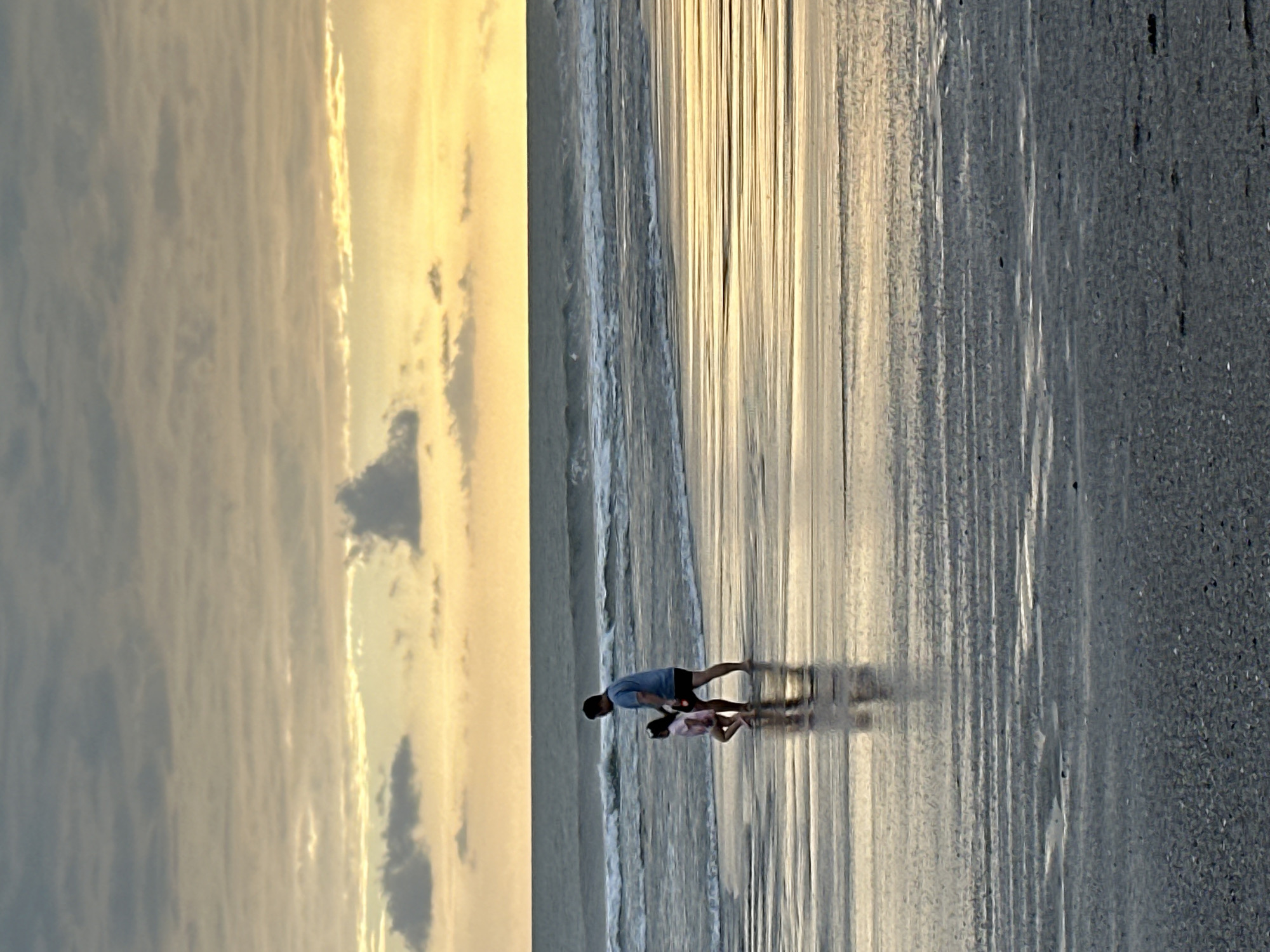 A father and child walk along the beach in North Kuta, Indonesia. The sky is filled with clouds in shades of orange and gray, indicating either sunrise or sunset. The ocean is calm with gentle waves, and the wet sand reflects the warm light from the sky. The father is wearing a blue t-shirt and black shorts, while the child is wearing a pink dress. They are walking towards the right of the frame, leaving footprints in the sand. The overall scene is peaceful and serene.