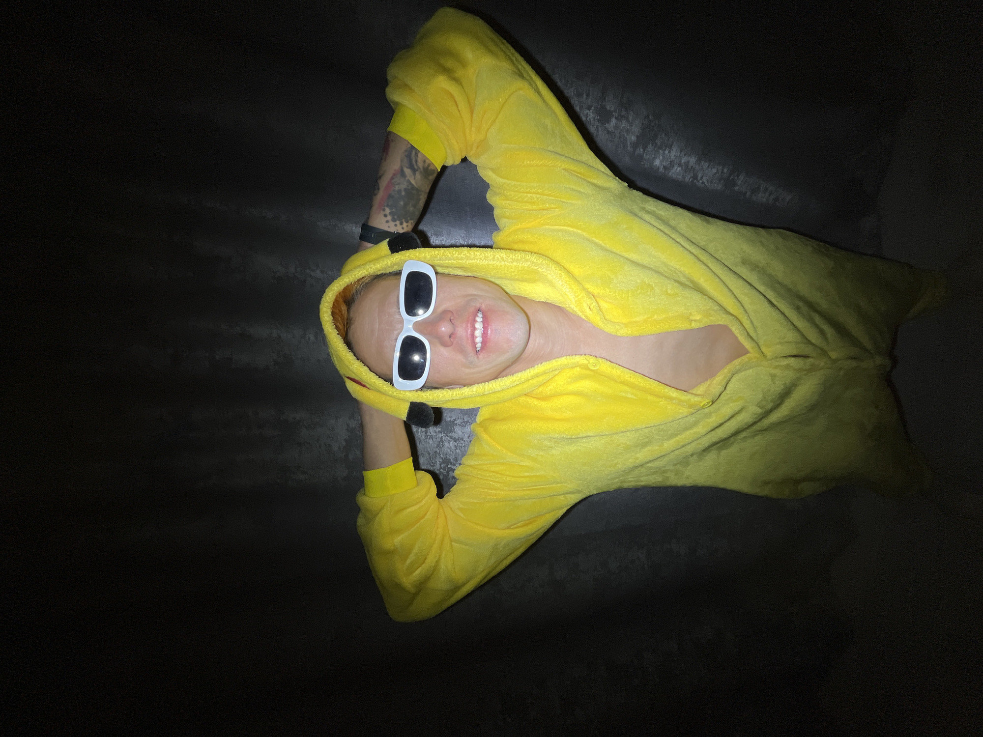 A person wearing a yellow Pikachu onesie and white sunglasses is lying down with their hands behind their head. The person is smiling and has visible tattoos on their arm. The location is not identifiable from the image.