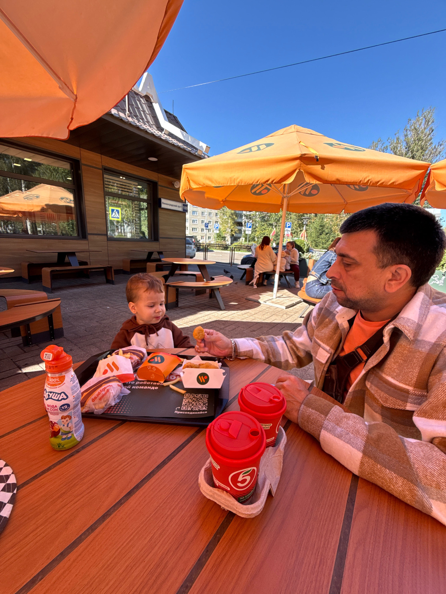 A man and a young child are seated at a wooden table outside a McDonald's restaurant in Rusanovo, Russia. The man, wearing a plaid shirt over an orange t-shirt, is holding a chicken nugget and appears to be feeding the child. The child, dressed in a brown hooded outfit, is looking at the nugget. On the table are food items, including burgers, fries, and drinks, served on a black tray. Two red paper cups with lids are placed in a holder. In the background, other diners are visible, and the McDonald's building with its signage and windows is present. The scene is illuminated by sunlight under a large orange umbrella.