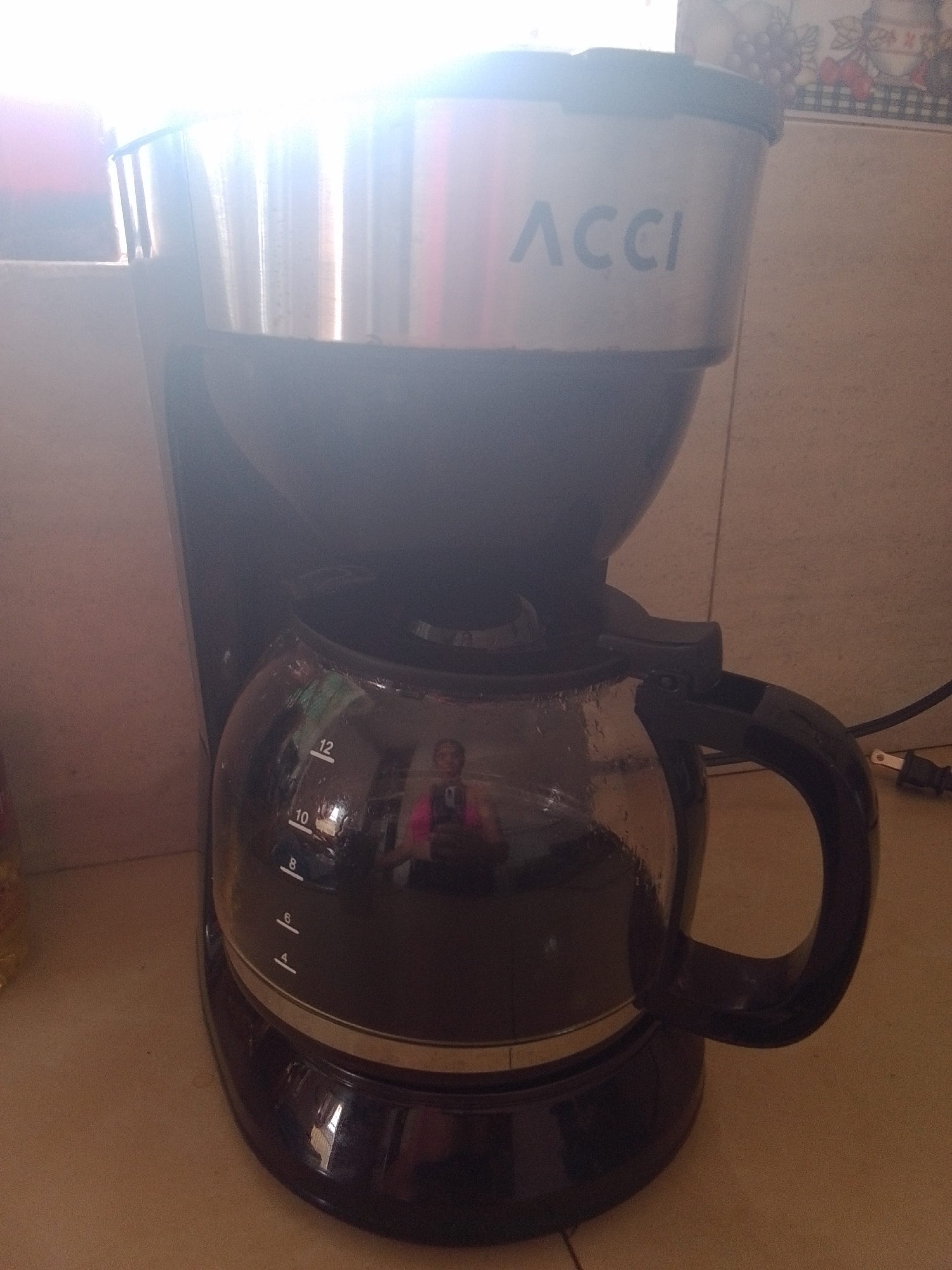 This is a medium-shot, eye-level photograph of a black and silver coffee maker positioned on a light-colored countertop. The coffee maker has a silver top portion with the brand name "ACCI" visible in blue lettering. Below this is a black reservoir that funnels into a glass carafe. The carafe has measurement markings on its side, with numbers 4, 6, 8, 10, and 12 clearly visible. The carafe appears to contain some liquid, and a reflection of a person holding a smartphone and wearing a pink shirt is visible on its surface. The background consists of a tiled wall, suggesting an indoor setting, possibly a kitchen. To the right of the coffee maker, a black electrical cord with a plug is visible, indicating the appliance is plugged in. The lighting in the image is bright, with a strong light source coming from the upper left, creating some glare and lens flare on the metallic parts of the coffee maker. The scene is static, with no discernible activity taking place beyond the implied function of the coffee maker. The overall impression is a practical, everyday object in a domestic environment. The location context is La Tagua, Venezuela.