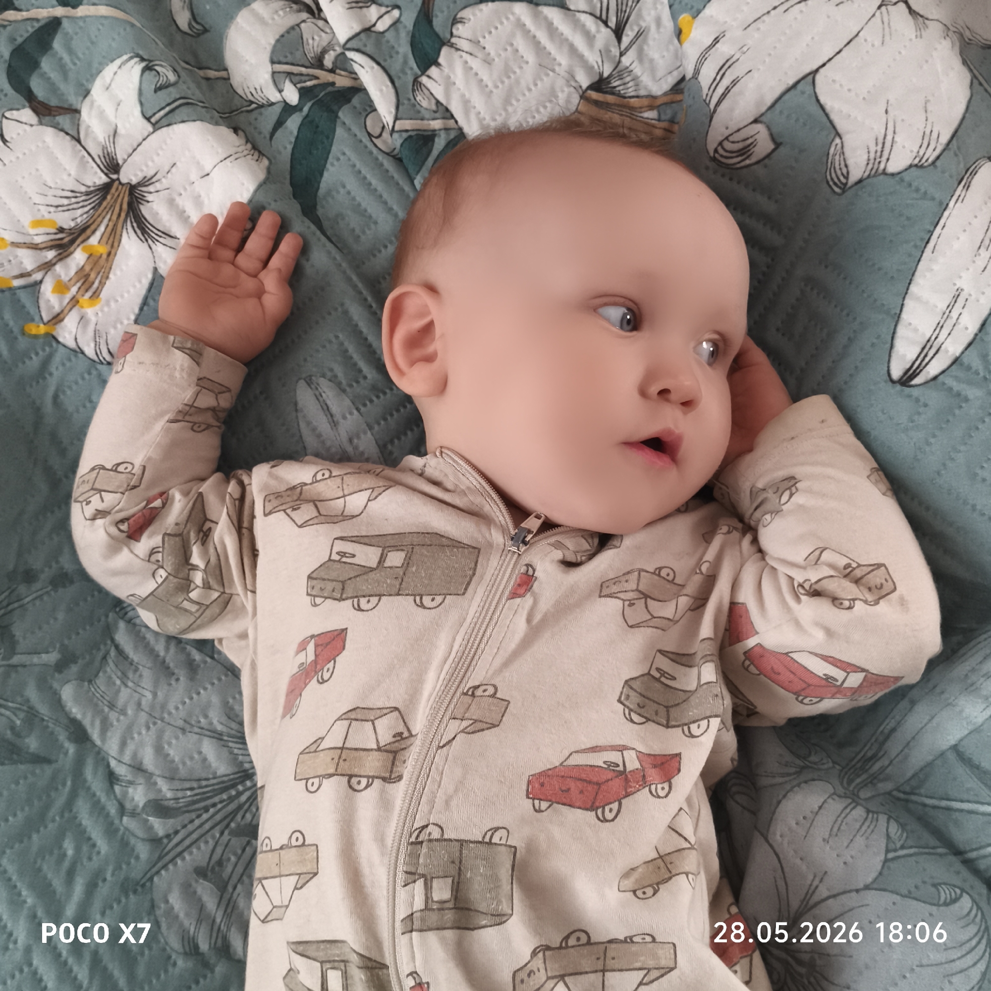 A baby lies on a floral-patterned quilt. The baby wears a car-patterned romper. The baby's arms are raised. The photo includes the text "POCO X7" and "28.05.2026 18:06". The location is Unknown, Unknown.