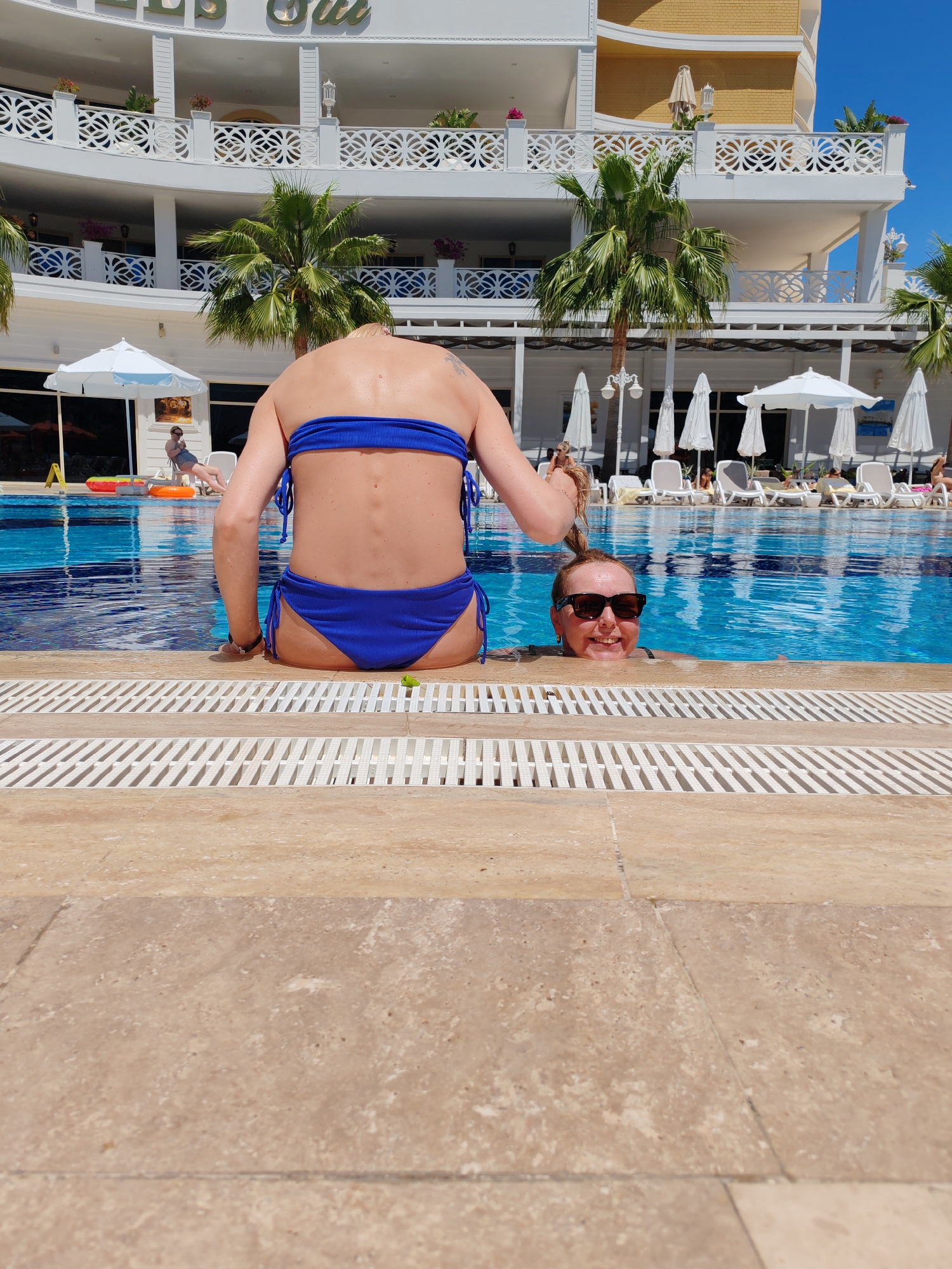 The image shows two women at the edge of a swimming pool. One woman is sitting on the edge, facing away from the camera, wearing a blue two-piece swimsuit. The other woman is in the pool, wearing sunglasses and smiling. The pool is surrounded by white lounge chairs and umbrellas. In the background is a multi-story white building with balconies and palm trees. This location is Alanya, Turkey.