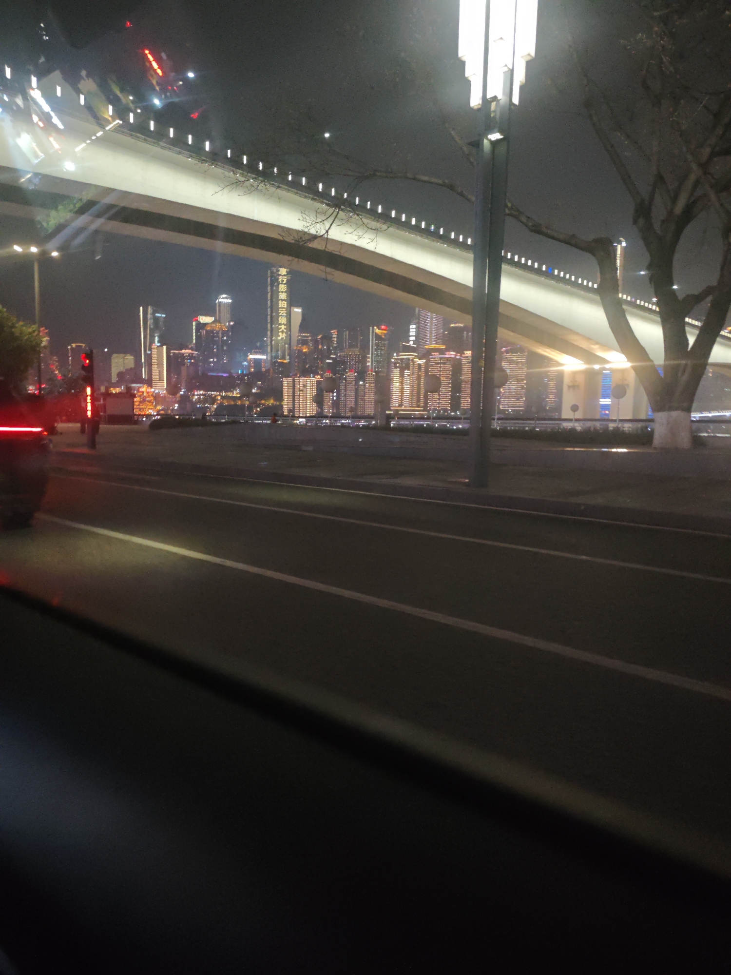 A nighttime view from a vehicle in 江北城街道, China. The road has multiple painted white lines. A car with illuminated tail lights is in the left side of the frame. A bridge with lights and support structures spans over a body of water. On the opposite shore is a cityscape with many illuminated buildings. A tall lamppost is visible on the right side. A bare tree is next to the lamppost.