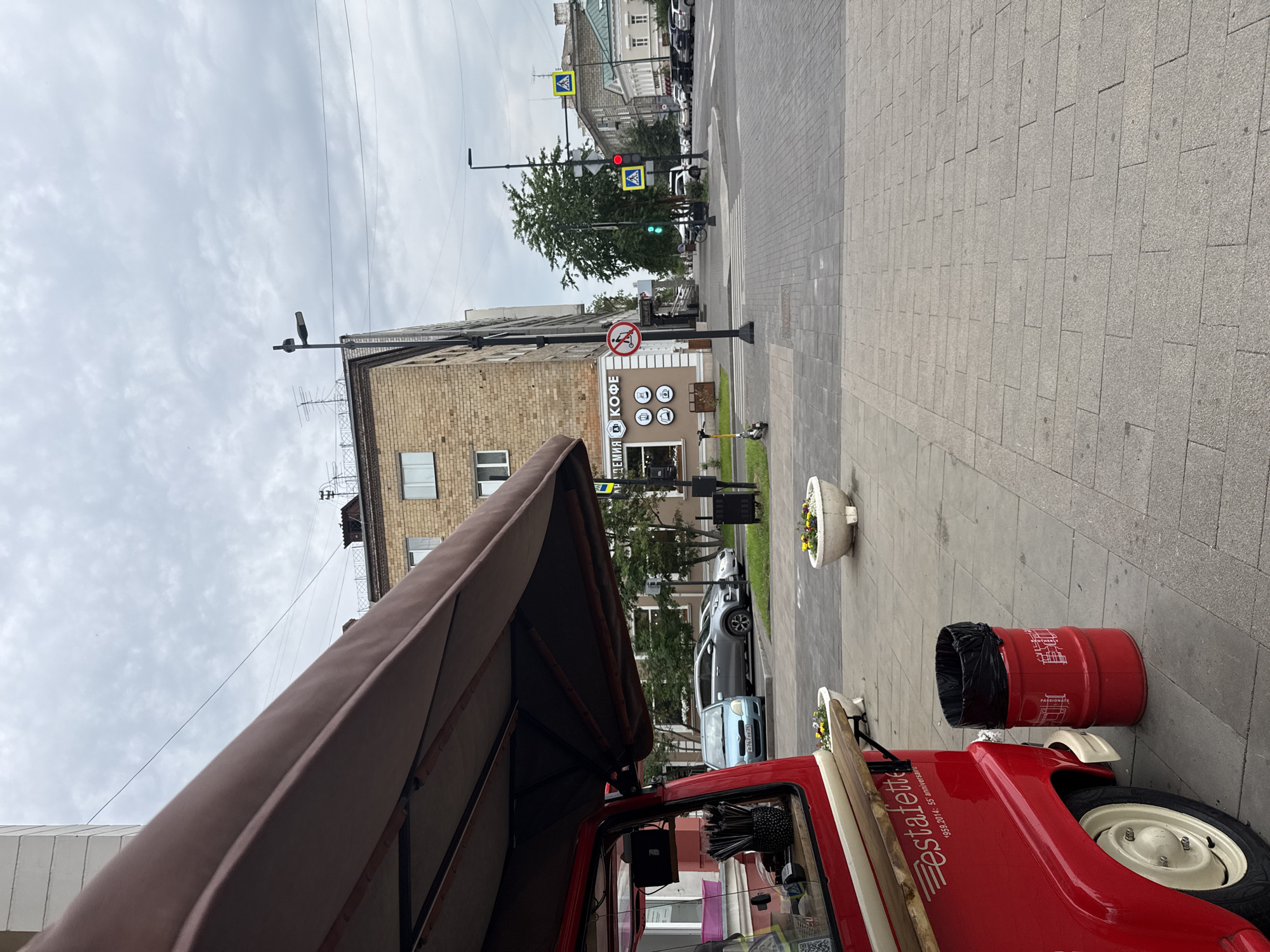 The image depicts a street scene in Krasnoyarsk, Russia. In the foreground, a red food cart with a brown awning is partially visible on the left. The cart has "estafette" written on its side. Next to the cart is a red cylindrical trash can with a black liner. The ground is paved with light gray bricks.
In the middle ground, a sidewalk runs along the street, with two white planters containing flowers. A yellow kick scooter is parked on the sidewalk. Further down the sidewalk, there is a building with signage that reads "АКАДЕМИЯ + КОФЕ" (Academy + Coffee). A traffic light is visible, showing a green light. A pedestrian crossing is also present.
The background shows a street with several parked cars and a multi-story building. The sky is overcast with gray clouds. The scene appears to be a typical urban environment on a cloudy day.