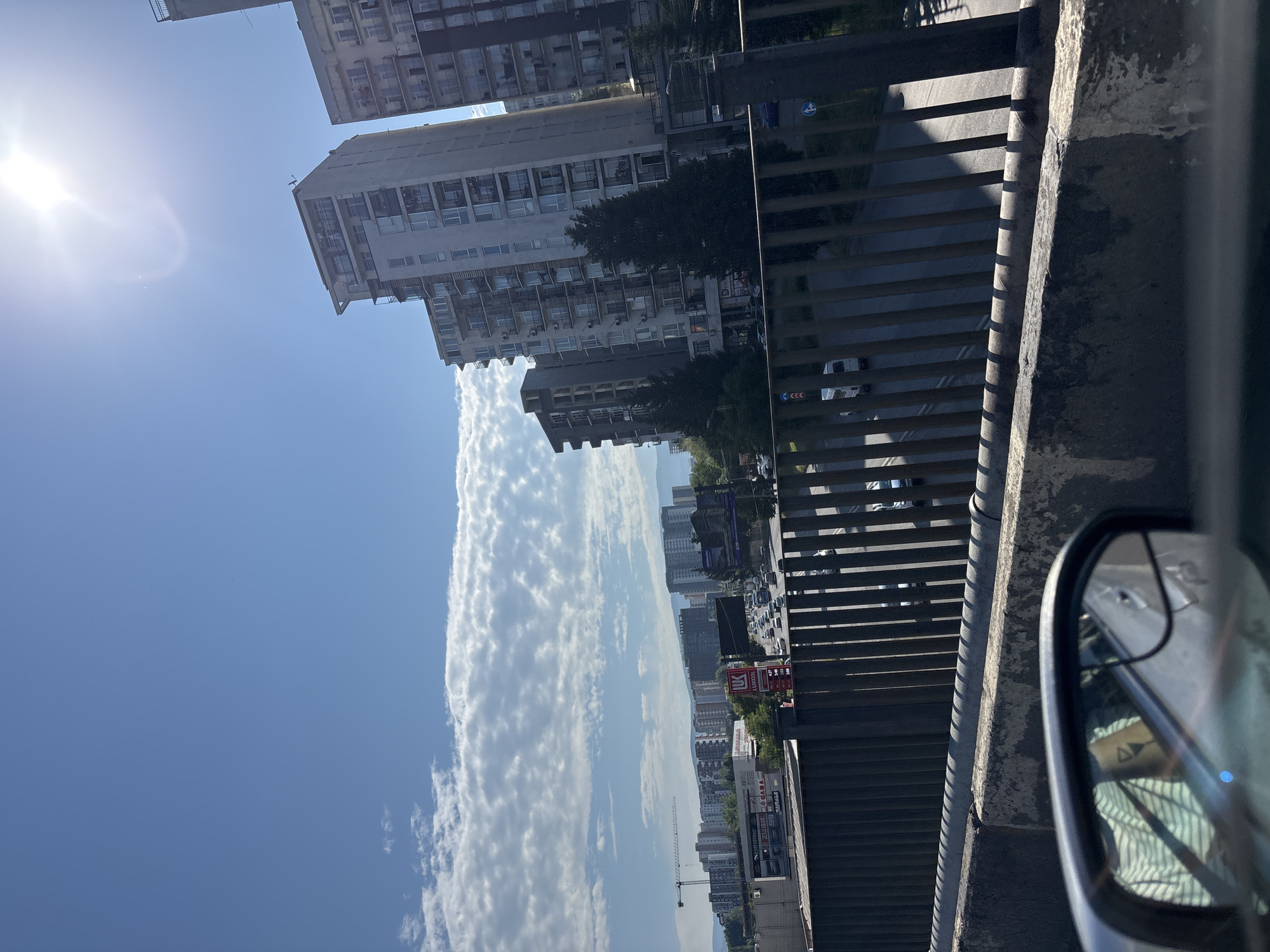 The image captures a view of a city in Tbilisi, Georgia, from an elevated position, likely a bridge or overpass. The scene is dominated by a bright, sunny sky with scattered white clouds. Several modern, multi-story apartment buildings are visible in the background, characterized by their rectangular shapes, numerous balconies, and light-colored facades. Some of these buildings appear to be under construction, with a crane visible in the distance. A busy road with a moderate amount of traffic, including cars and a white van, runs below the overpass. A Lukoil gas station sign is prominently displayed on the side of the road. The foreground features a concrete barrier and a metal railing. A car's side-view mirror is visible in the bottom left corner, reflecting the interior of the car. The overall impression is of an urban environment on a clear, sunny day.