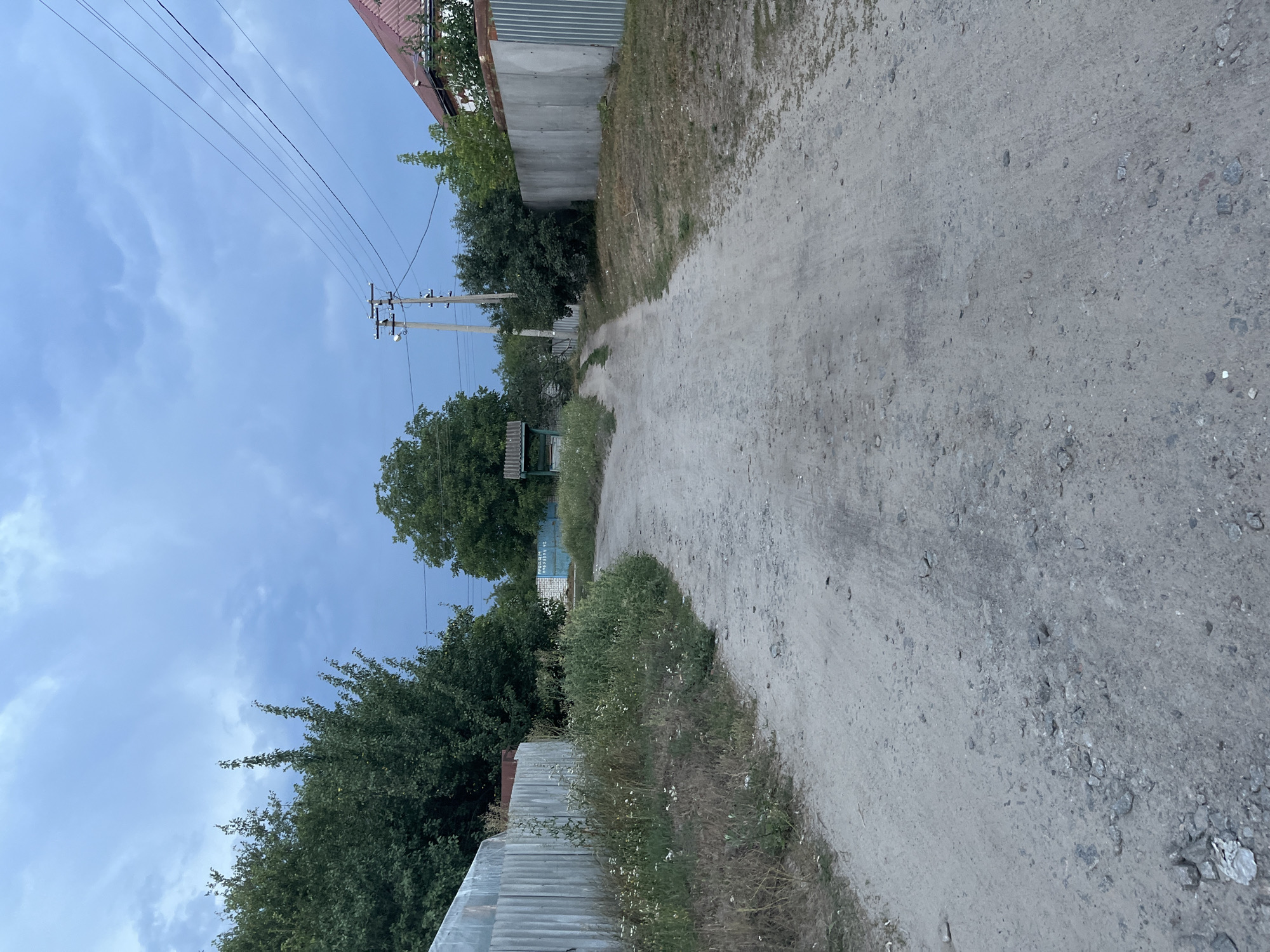 The image depicts a dirt road in Vvedenka, Ukraine. The road is flanked by corrugated metal fences on both sides. To the left, a fence is partially obscured by overgrown grass and wildflowers. To the right, a similar fence runs along the road, with a house with a red roof visible in the background. Trees and power lines are present throughout the scene. The sky is cloudy and appears to be overcast. In the distance, a simple wooden structure with a roof, possibly a well or a shelter, can be seen on the left side of the road. There are no people or animals visible in the image. The overall impression is one of a rural or semi-rural setting.