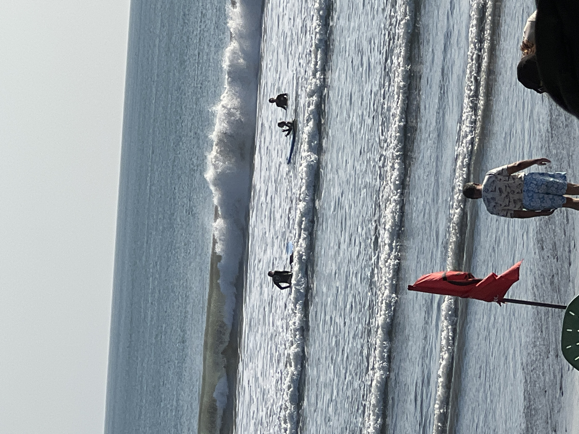 The image shows a beach scene with several surfers in the ocean. In the foreground, a man in a patterned shirt and blue shorts stands on the shore facing away from the camera. To his left, a red flag on a pole stands in the sand. In the middle ground, several surfers are in the water, with one sitting on a surfboard. The background shows the ocean with waves and a clear sky. The scene appears to be set in the city of Kuta, Indonesia, a well-known surfing destination.