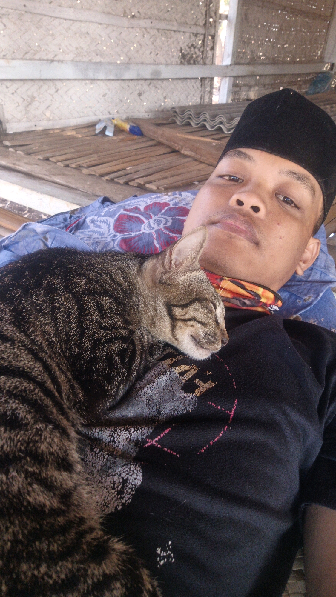 A close-up, eye-level shot captures a person lying down, seemingly on a bamboo-slatted surface, with a tabby cat asleep on their chest. The person is wearing a black taqiyah, a patterned blue and red cloth around their neck, and a black t-shirt with a faded graphic design. The cat is nestled comfortably, its eyes closed. The background shows a wall constructed of woven bamboo, suggesting an outdoor or semi-outdoor shelter. The lighting is bright and appears to be natural daylight. The overall scene conveys a sense of peace and relaxation. The location is identified as Sokaramme Timur, Indonesia.