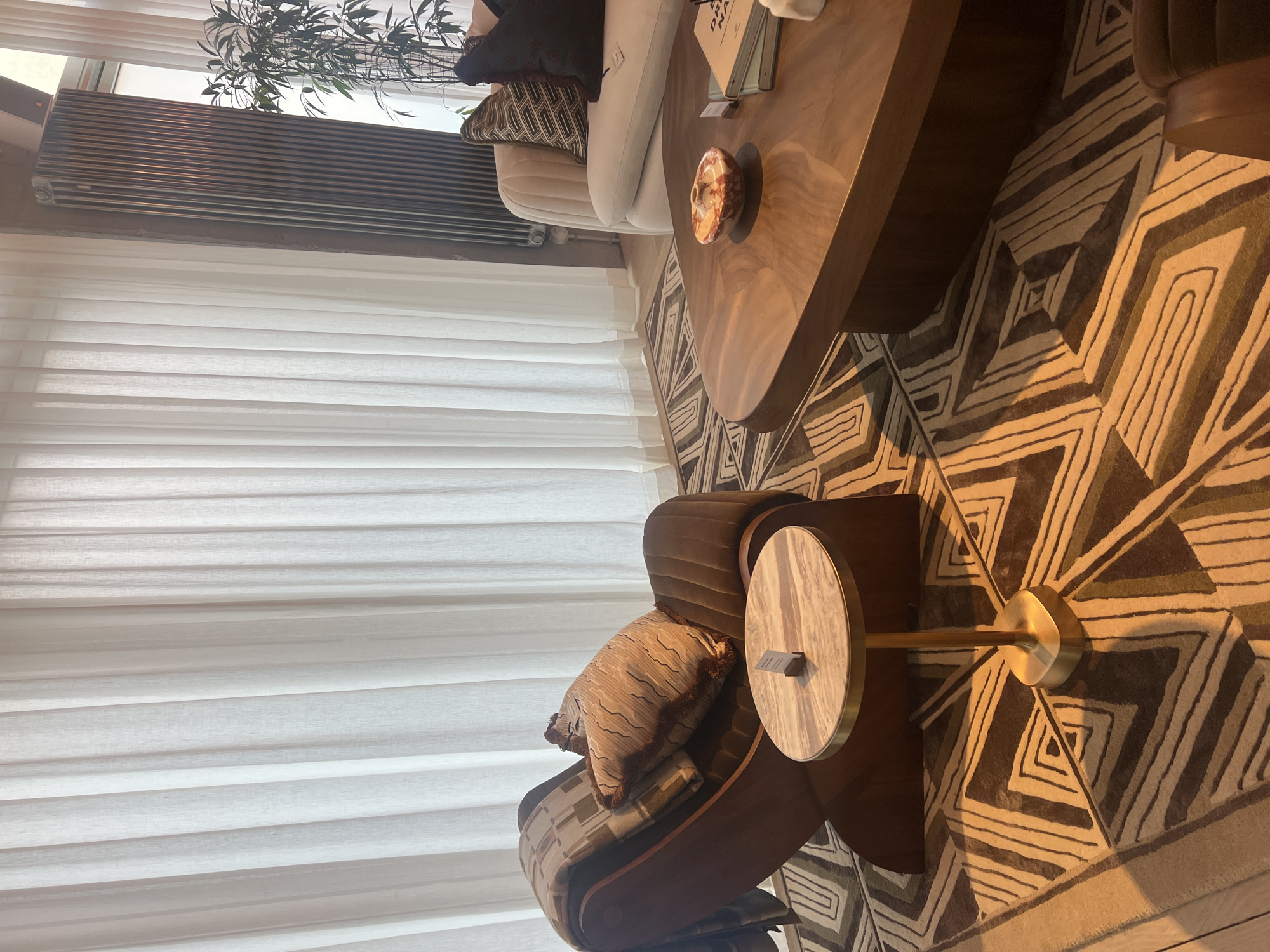 A stylish and well-appointed living space, likely a lounge or living room, is captured in this image, rotated 90 degrees clockwise. Correctly oriented, the left side of the frame features large windows covered by long, sheer, off-white curtains that diffuse natural light. Below the windows, a dark, horizontal radiator or heating unit is mounted.
In the center, a cream or light beige sofa is adorned with dark and geometric-patterned throw pillows. In front of the sofa, a large, dark wooden coffee table with a unique, organic shape holds a small, marbled decorative object and a stack of books or magazines. Partially visible text on the books reads "DREAM NA" or similar. The floor is covered by a distinctive rug featuring a bold geometric pattern of interlocking triangles and squares in shades of brown, beige, and olive green.
To the right, a comfortable dark brown armchair, accented with a patterned cushion and a throw blanket, sits beside a small, elegant side table. This side table has a round, light-colored marble top supported by a slender brass or gold-toned stem and a square base. A small, dark card is placed on the side table, but its text is illegible. A green plant with long, thin leaves is partially visible in the upper left background, adding a natural element.
The room is illuminated by ample natural light, suggesting it is daytime. The decor exhibits a modern, sophisticated aesthetic with earthy tones and rich textures, creating a comfortable and inviting atmosphere. No people are present, indicating a tranquil scene. The setting suggests a contemporary interior, potentially located in Berlin, Germany.