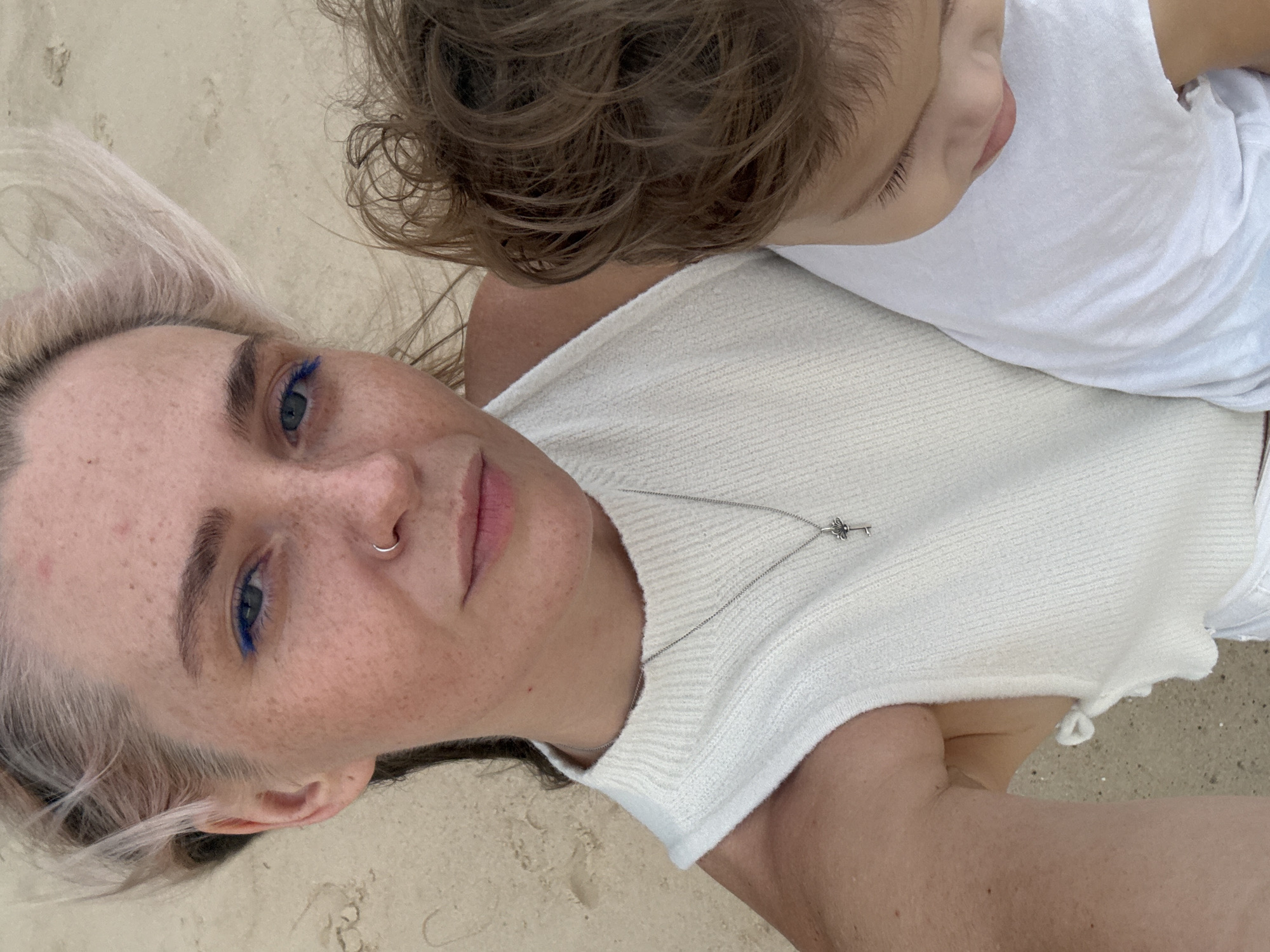 The image shows a close-up, high-angle selfie of a woman holding a child. The woman has light-colored hair, freckles, and blue eyeliner. She is wearing a white sleeveless top and a silver necklace with a key pendant. The child, partially visible, has brown hair and is wearing a white t-shirt. They appear to be on a sandy surface, suggesting an outdoor setting like a beach. The lighting indicates daylight.