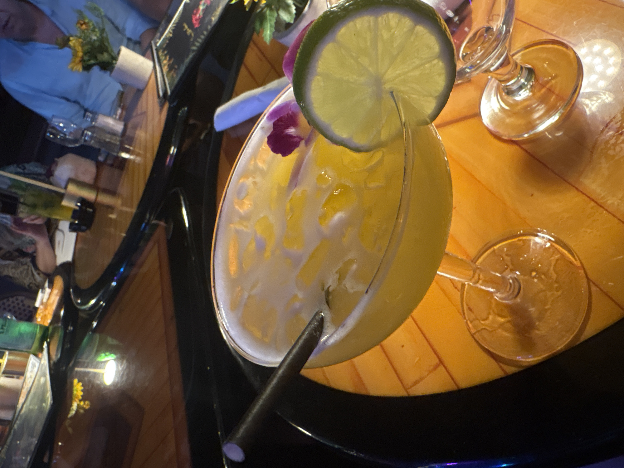 A vibrant, yellow cocktail in a martini-style glass dominates the center of this diagonally oriented image. The drink, filled with ice and possibly fruit pulp, is garnished with a fresh lime wheel on the rim and a small, delicate purple orchid flower floating on its surface. A dark, possibly metal, straw is submerged in the cocktail.
The glass rests on a glossy, warm-toned wooden table that reflects ambient lighting, indicating an indoor setting, likely a bar or restaurant, during the evening or night. The table surface shows a subtle patterned texture, possibly wood grain or an inlay design. Scattered around the main cocktail are other objects: two empty stemmed glasses, one upright and one on its side; a small white pot containing green foliage and yellow artificial flowers; and a dark, rectangular object, possibly a menu holder or framed artwork, displaying indistinct colorful patterns. A green bottle in a holder is also partially visible on the left side of the table.
In the blurred background, fragments of two individuals can be seen. One person is wearing a light blue shirt, while another person's arm and a patterned garment are visible. The scene suggests a casual social gathering where people are enjoying drinks. The tropical flower garnish and vibrant cocktail color are consistent with the provided location context of Miami Beach. No clearly legible text is discernible in the image.