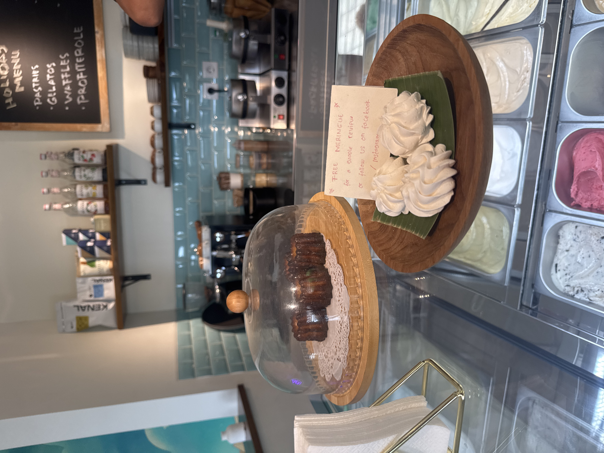 The image shows a display case filled with various flavors of ice cream. In the foreground, there are three white meringues on a wooden plate lined with a green leaf. A small handwritten note is placed beside the meringues, offering "FREE MERINGUE for a google review or follow us on facebook / instagram." Above the ice cream display, there are two cannelés under a glass dome. In the background, shelves hold bottles of syrup, packets of coffee or tea, and waffle makers. A chalkboard menu is visible on the right side of the image, listing "HOLIDAY MENU" with items such as "PASTRIPS", "GELATOS", "WAFFLES", and "PROFITEROLLE". The establishment appears to be a cafe or dessert shop. There is no indication of the city of Abang, Indonesia in the image.