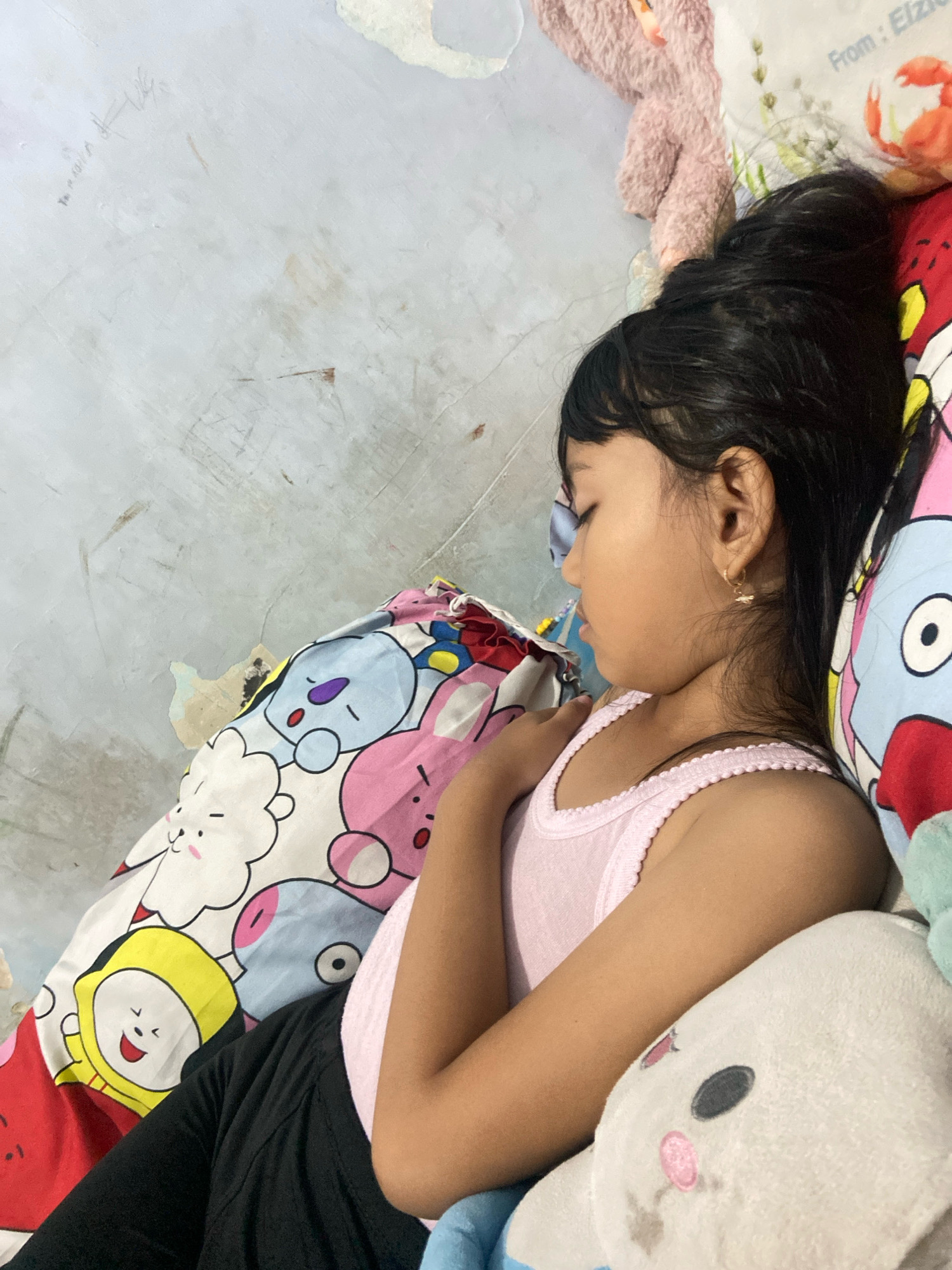 A young girl sleeps soundly in bed, her head resting on a pillow adorned with cartoon characters. She is wearing a pink tank top and black pants. A plush toy is nestled beside her. The scene is indoors, possibly a bedroom, with a wall that appears textured and slightly worn in the background. The lighting suggests it could be daytime, though the specific time is not clear. The atmosphere is peaceful and suggests a moment of rest. There is no visible text in the image. The context provided indicates the location as Gelam, Indonesia.