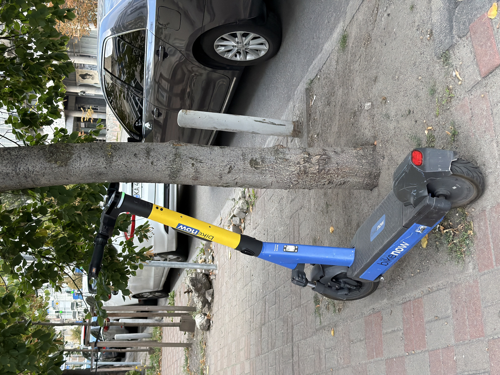 An electric scooter with "BikeNOW" branding in yellow and blue is parked on a sidewalk next to a tree. The scooter is in the foreground, angled slightly away from the viewer. Behind the scooter, a dark-colored sedan is parked along the street. The sidewalk is paved with brick. In the background, other parked cars and buildings are visible, along with trees and streetlights, suggesting an urban environment in Kyiv, Ukraine.