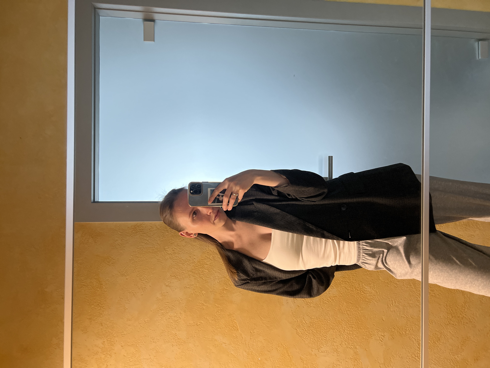 A woman is taking a selfie in a bathroom mirror. She is wearing a white top, gray sweatpants, and a dark gray blazer. The bathroom has yellow textured walls and a large mirror with a white frame. The background reflected in the mirror is blurred, but appears to be an outdoor scene, possibly a bright sky. There is no discernible evidence to identify the location as Khimki, Russia.